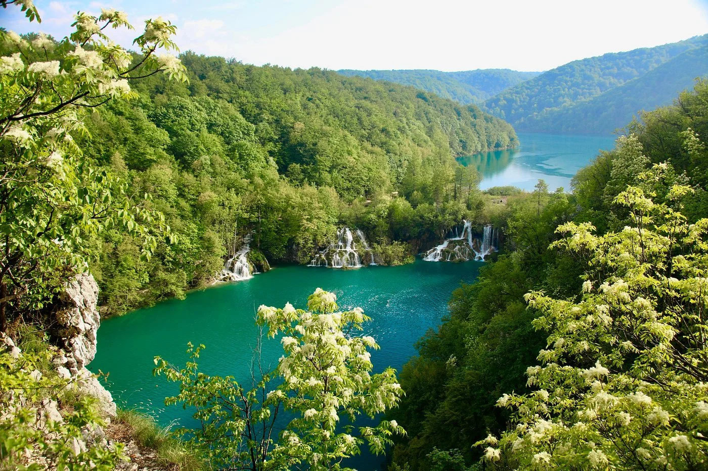 A scenic view of a river or lake surrounded by lush green trees and hills, with small waterfalls flowing into the water.