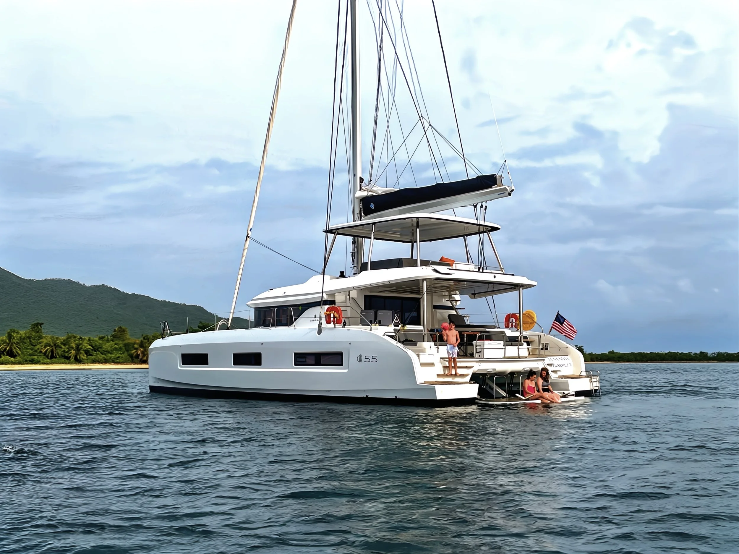 A white sailboat with three levels on the water, with three people relaxing on the back platform and deck, and the American flag at the stern. Land with green trees and hills in the background, under a partly cloudy sky.