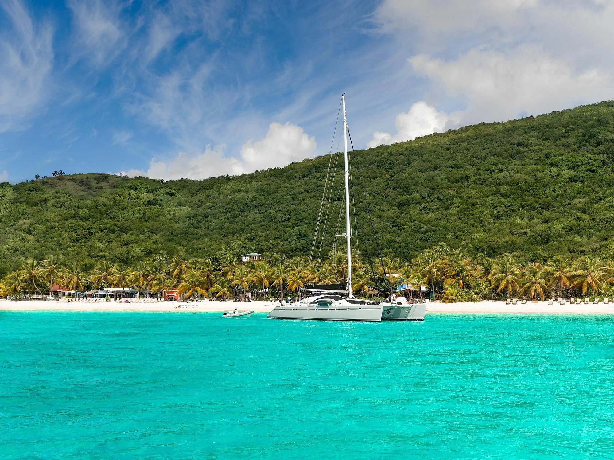 A sailboat anchored in clear, turquoise water near a tropical beach with palm trees and a lush green mountain in the background.