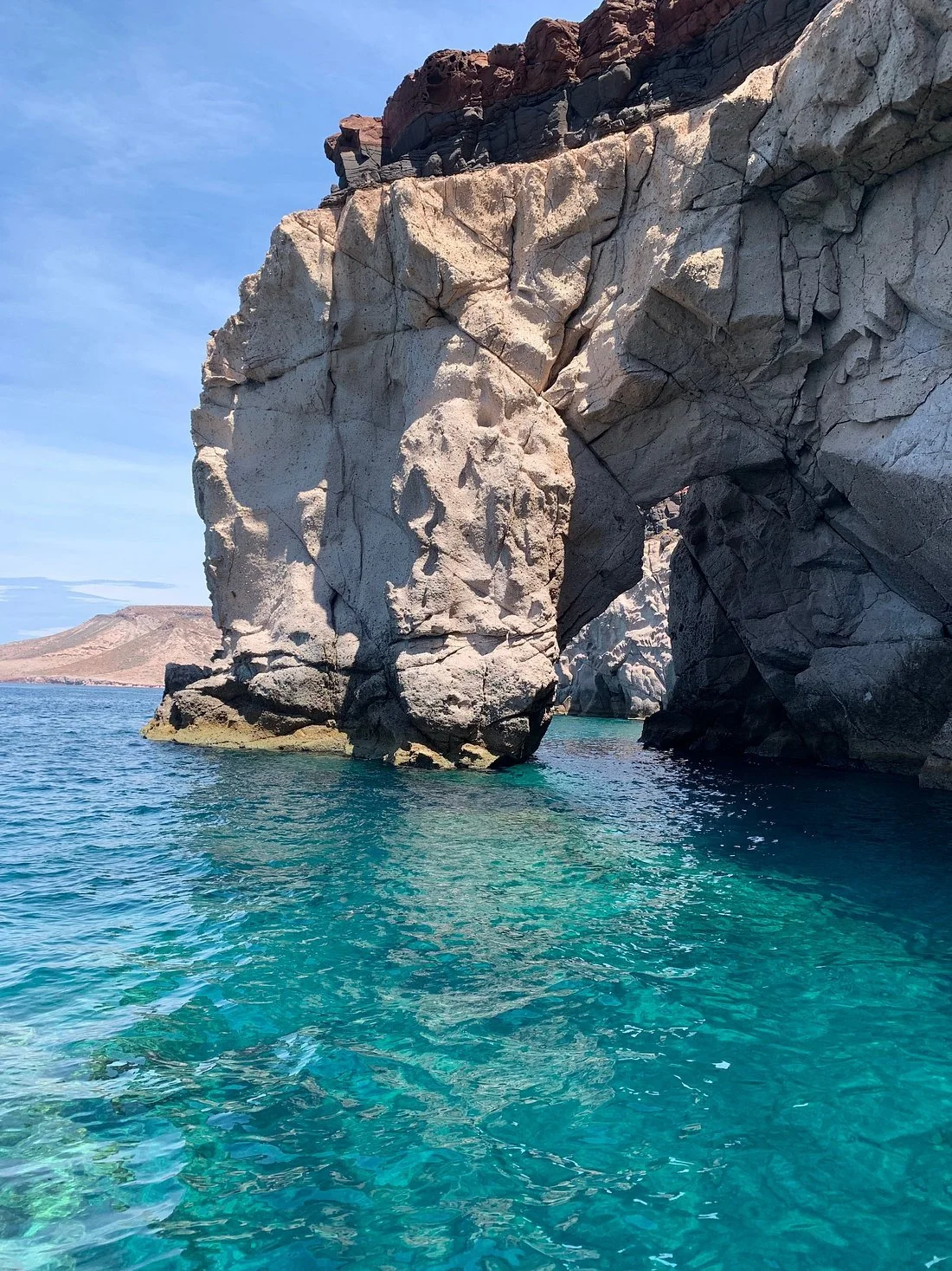 Large rock formation with an arch over turquoise water, landscape in the background, clear blue sky.