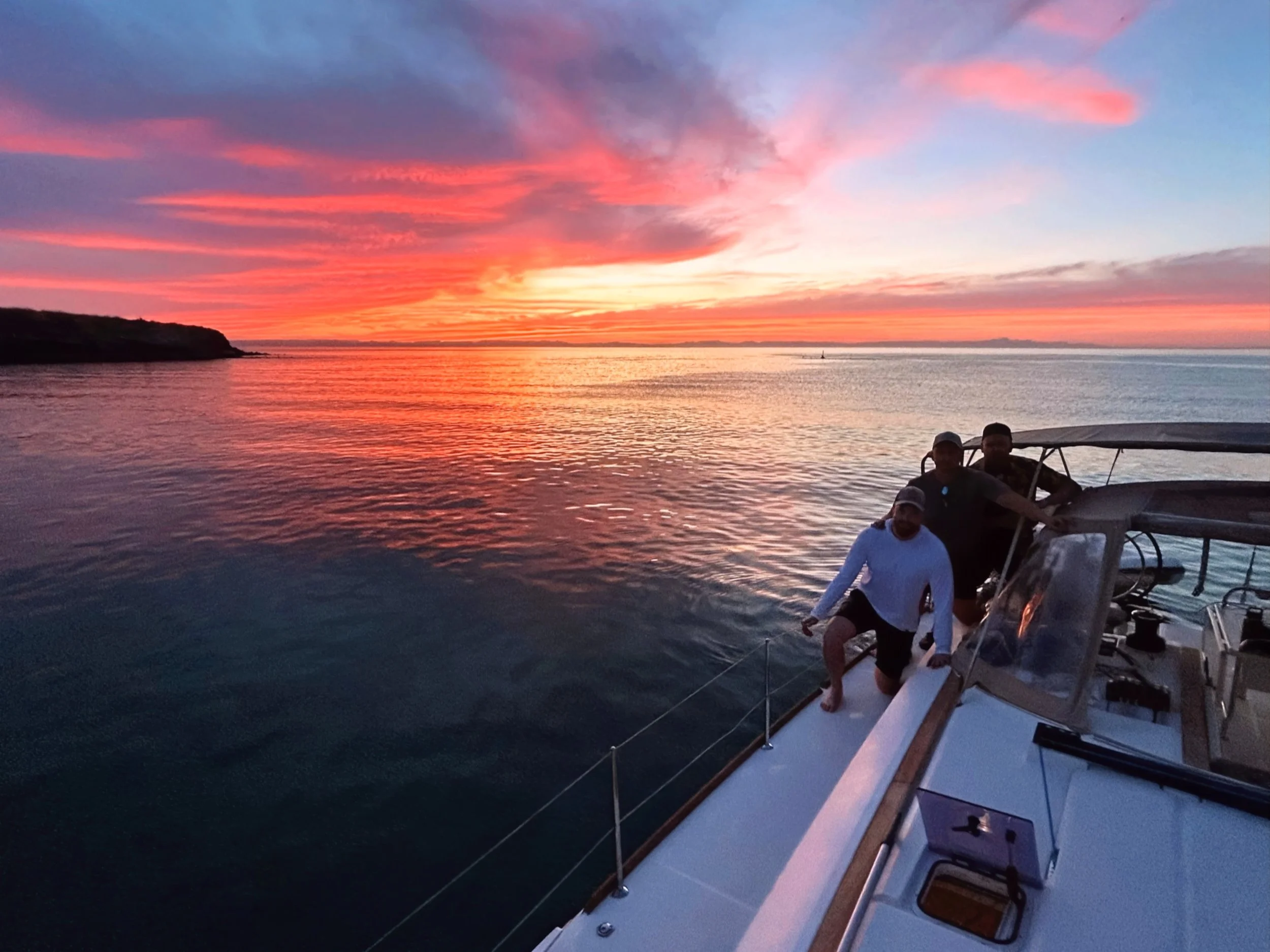 Three people on a sailboat at sunset, with a colorful sky and calm water in the background.