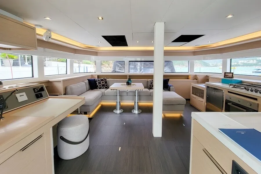 Modern yacht interior with large windows, L-shaped seating with cushions, a central table, and a kitchenette with stove and appliances.