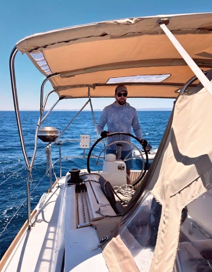 A man standing at the steering wheel of a boat on the open ocean under a clear blue sky.