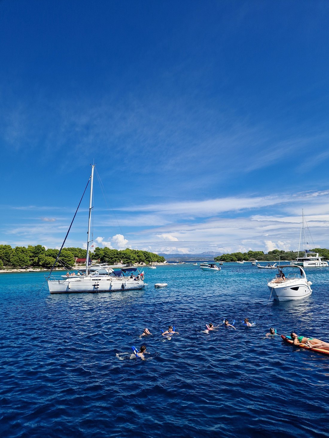 People snorkeling and relaxing on a raft in a clear blue body of water near sailboats and a lush green shoreline under a partly cloudy sky.