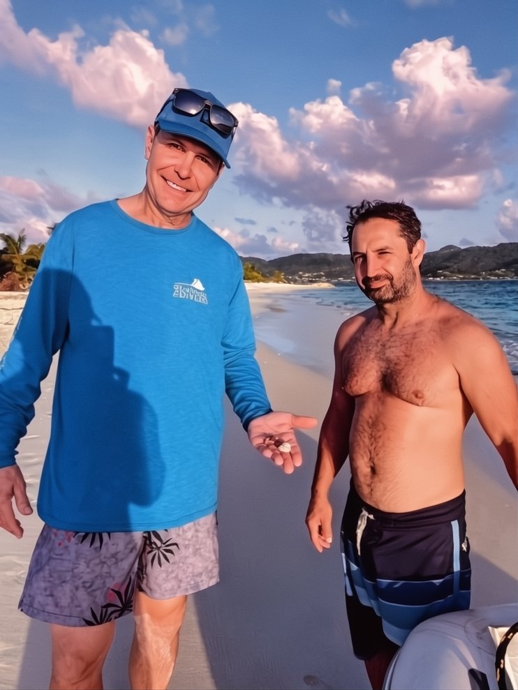 Two men on a beach, one wearing a blue slice, shorts, and sunglasses on his head, smiling and holding a shell, while the other is shirtless and wearing swim trunks, standing near the ocean with mountains and clouds in the background.