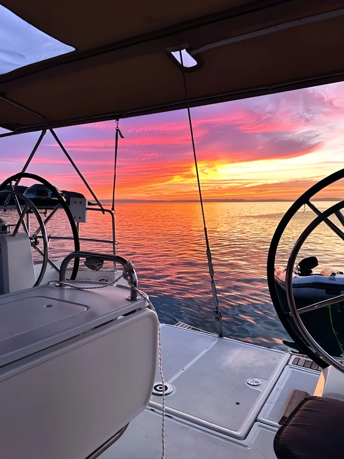 The photo shows the deck of a boat at sunset, with a colorful sky of pink, purple, and orange reflected on the water. The boat's steering wheels and part of the canopy are visible.