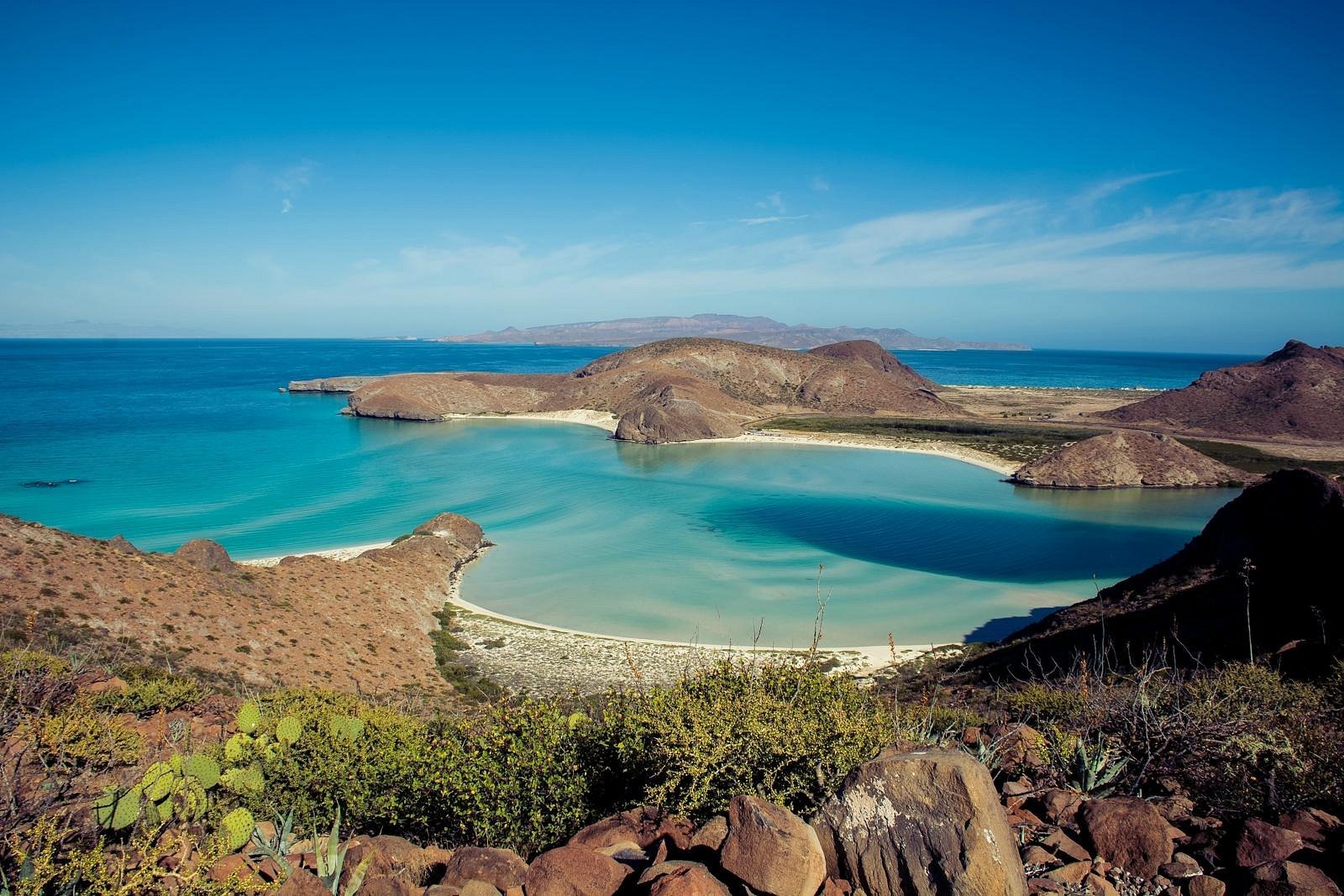A coastal landscape featuring a lagoon with turquoise water, surrounded by brown desert hills and mountains under a clear blue sky.