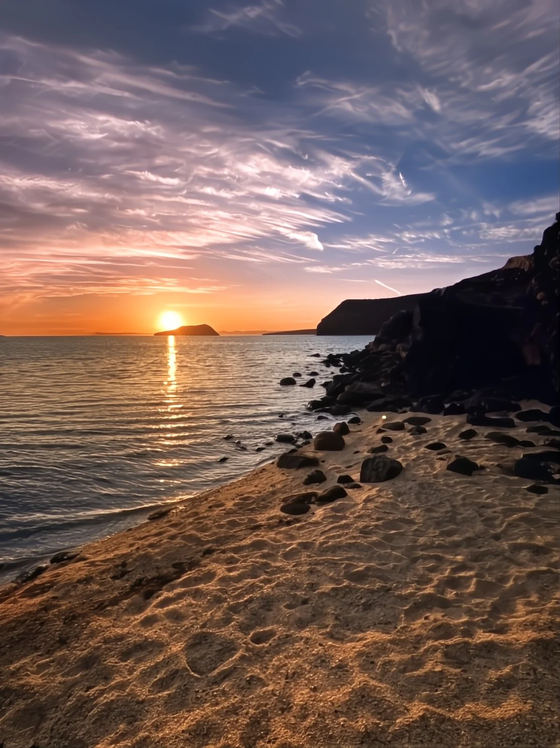 Sunset over the ocean with a rocky beach in the foreground, clouds in the sky, and an island on the horizon.