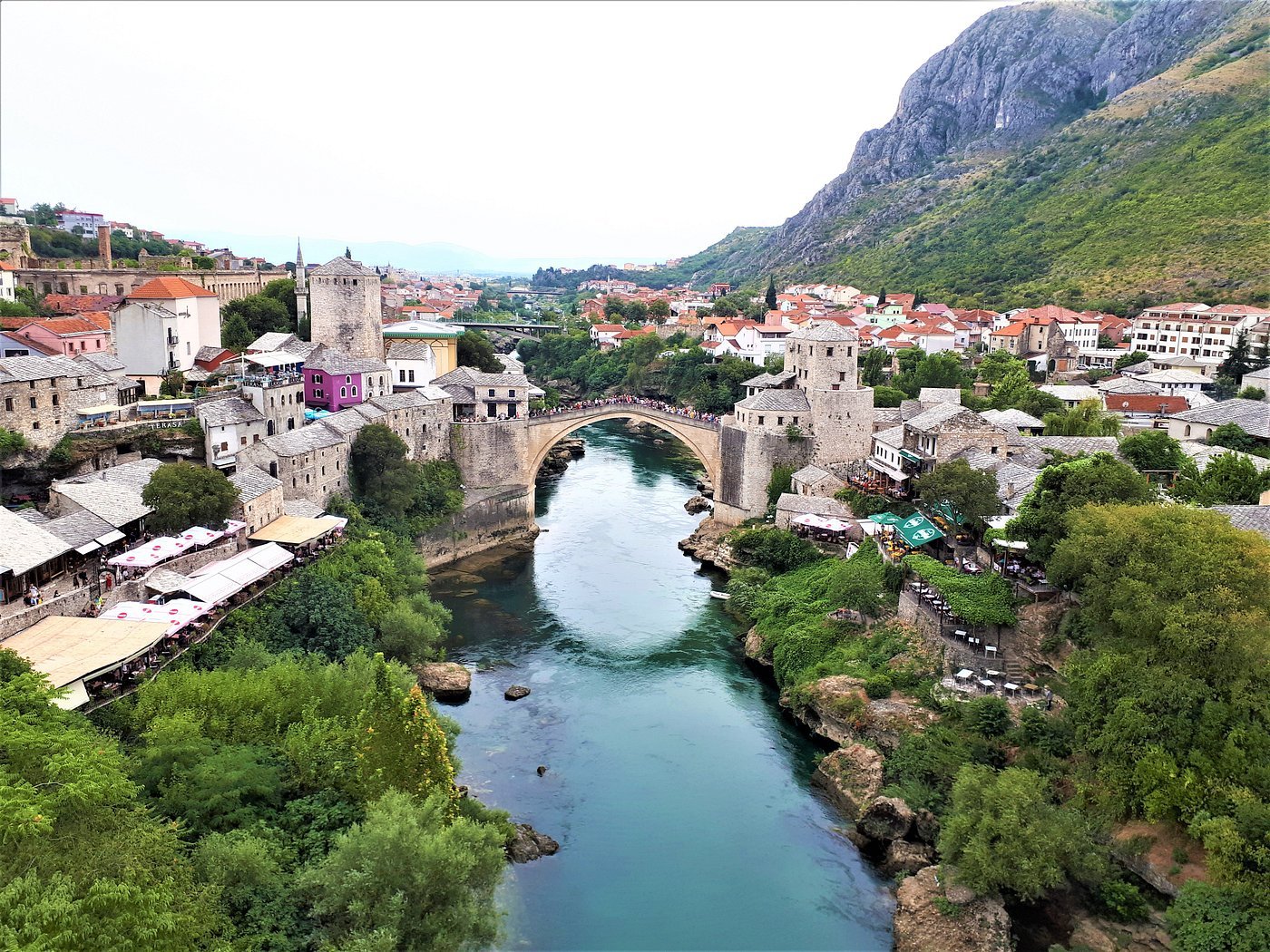 A scenic view of a historic European town with a river running through it, stone buildings, a bridge, green hills, and mountains in the background.