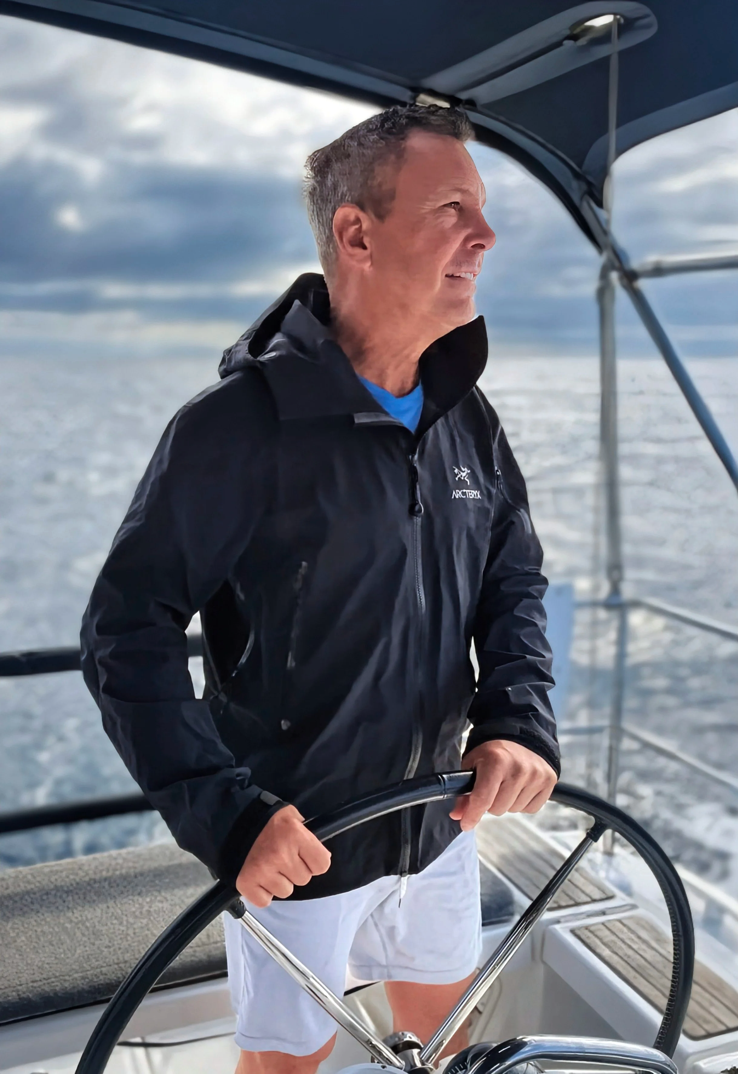 A man steering a boat at sea, wearing a black Arc'teryx jacket and white shorts, with cloudy sky background.