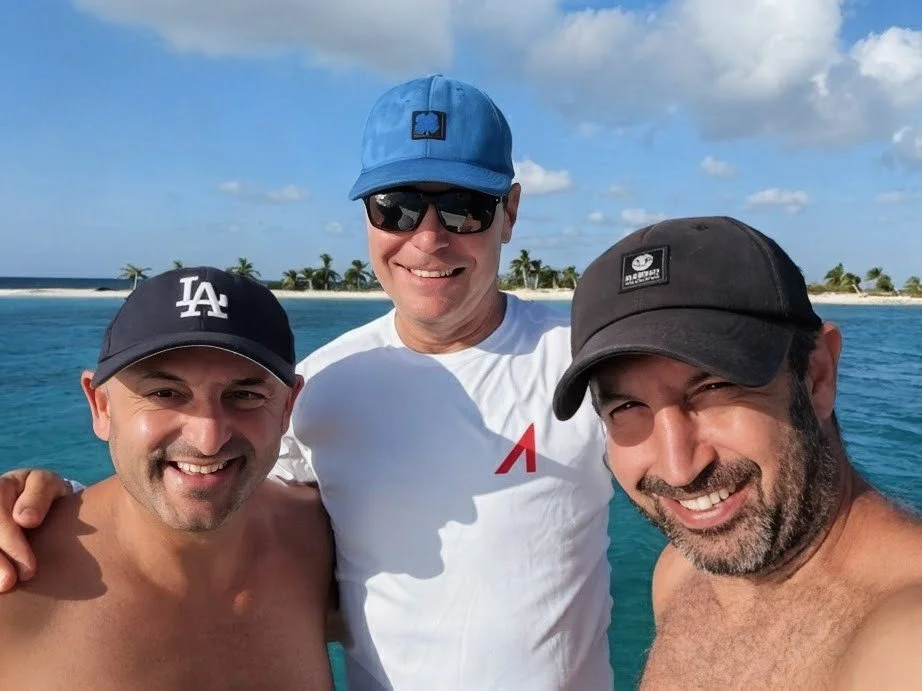 Three men in a tropical setting taking a selfie with clear blue water and palm trees in the background, all smiling.