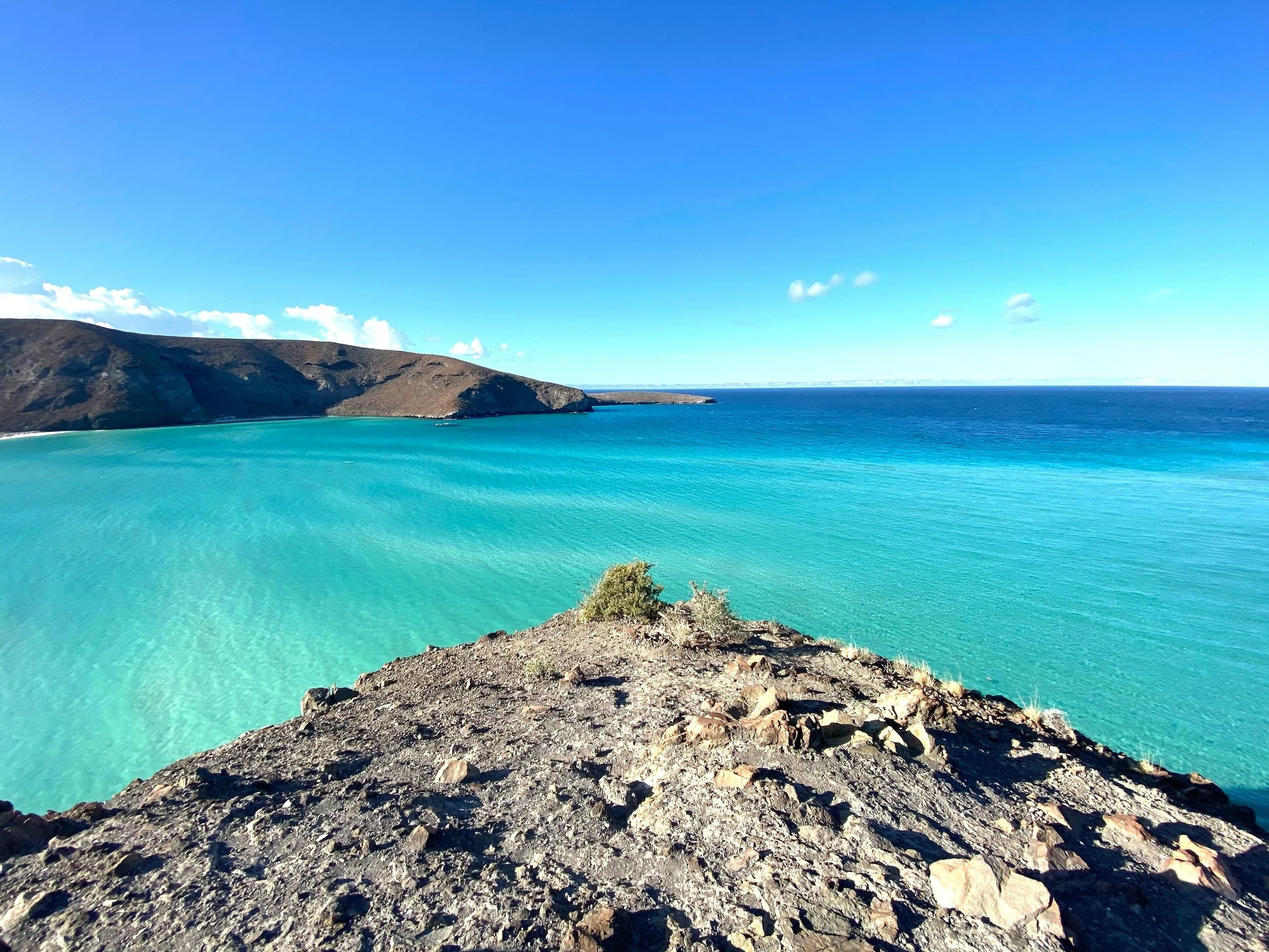 A scenic view of turquoise ocean waters meeting a rocky shoreline under a bright blue sky, with distant hills in the background.