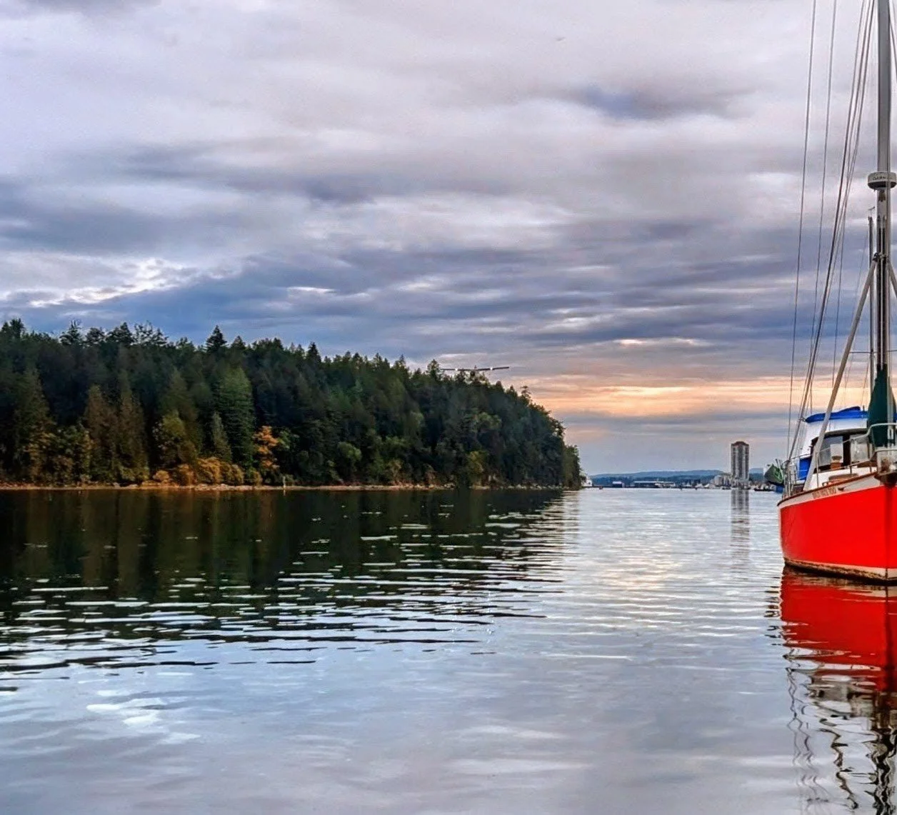 A calm river scene at dusk with a red sailboat docked on the right side, lush green forested shoreline on the left, and a cloudy sky with the sun setting in the distance.