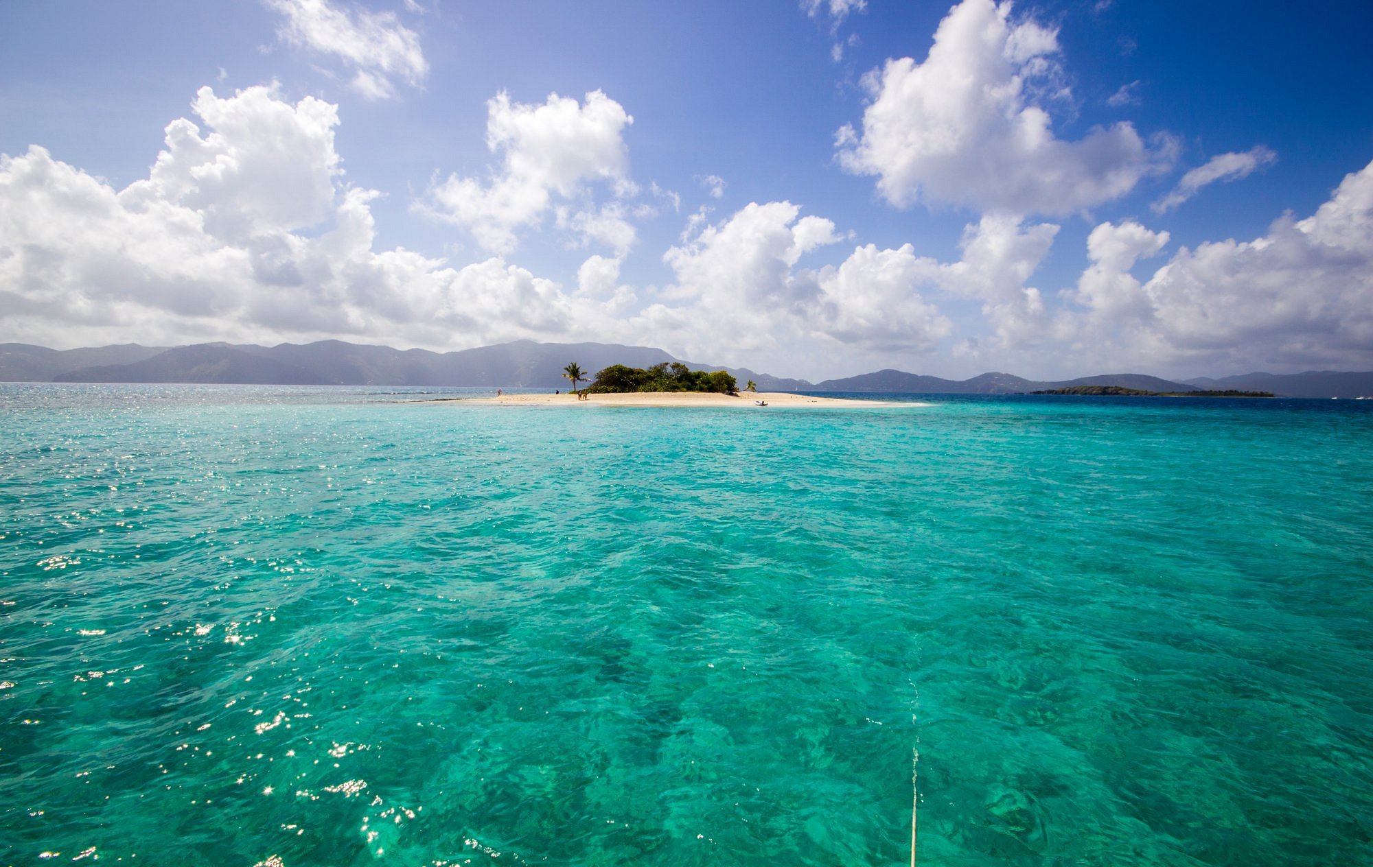 Tropical island with sandy beach, palm trees, and turquoise ocean waters under a partly cloudy sky, with mountains in the background.