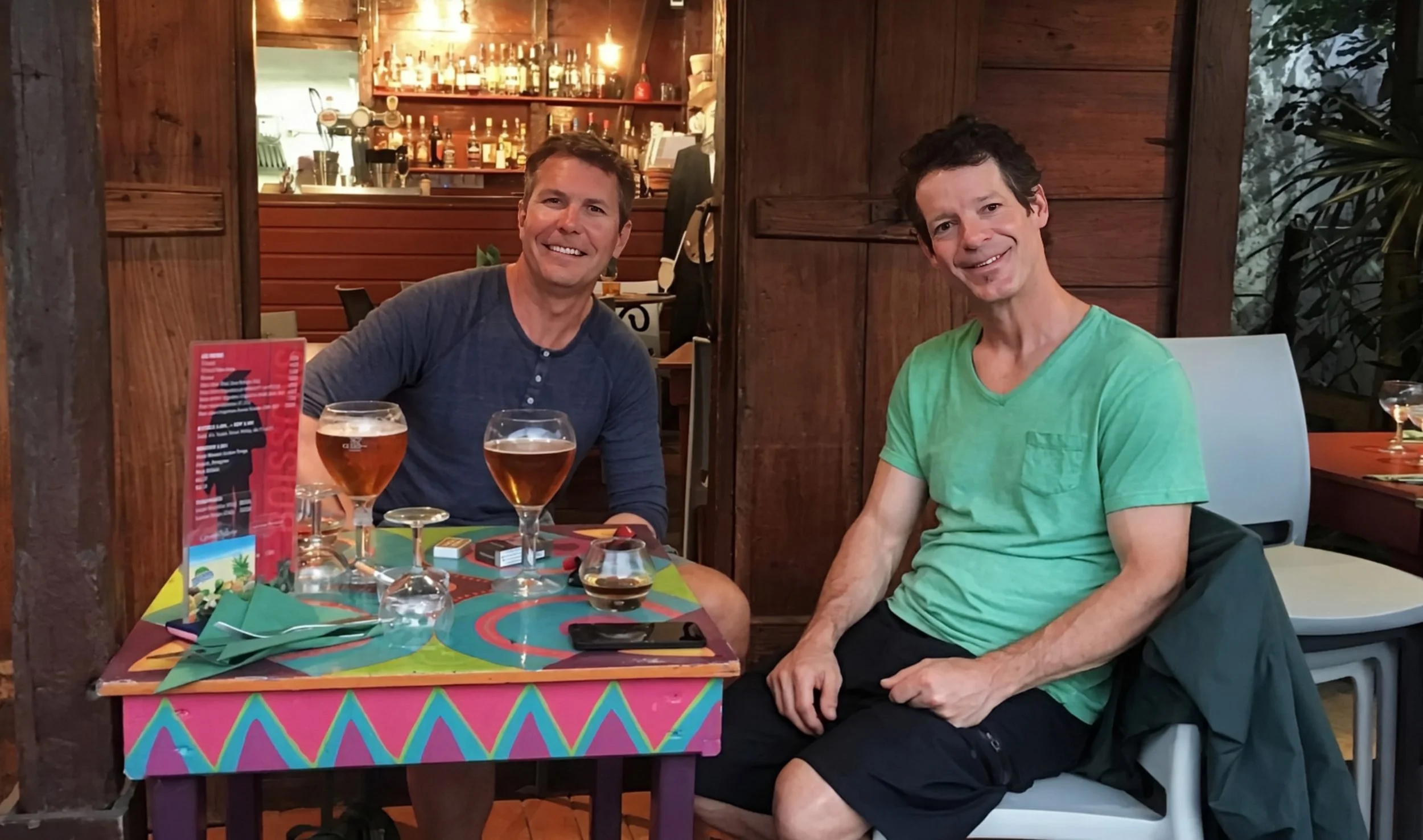 Two smiling men sitting at a colorful table with drinks in a restaurant, with a wooden interior and a bar in the background.