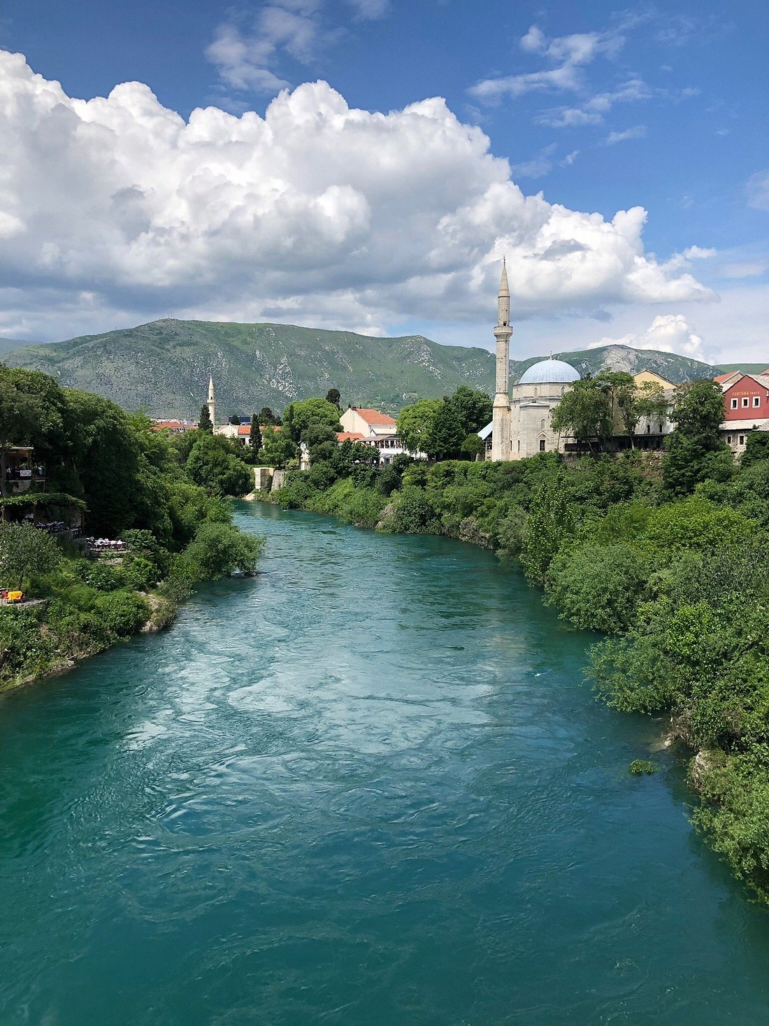 A river flowing through a lush green landscape with a small town and a mosque with a tall minaret, mountains in the background, under a partly cloudy sky.