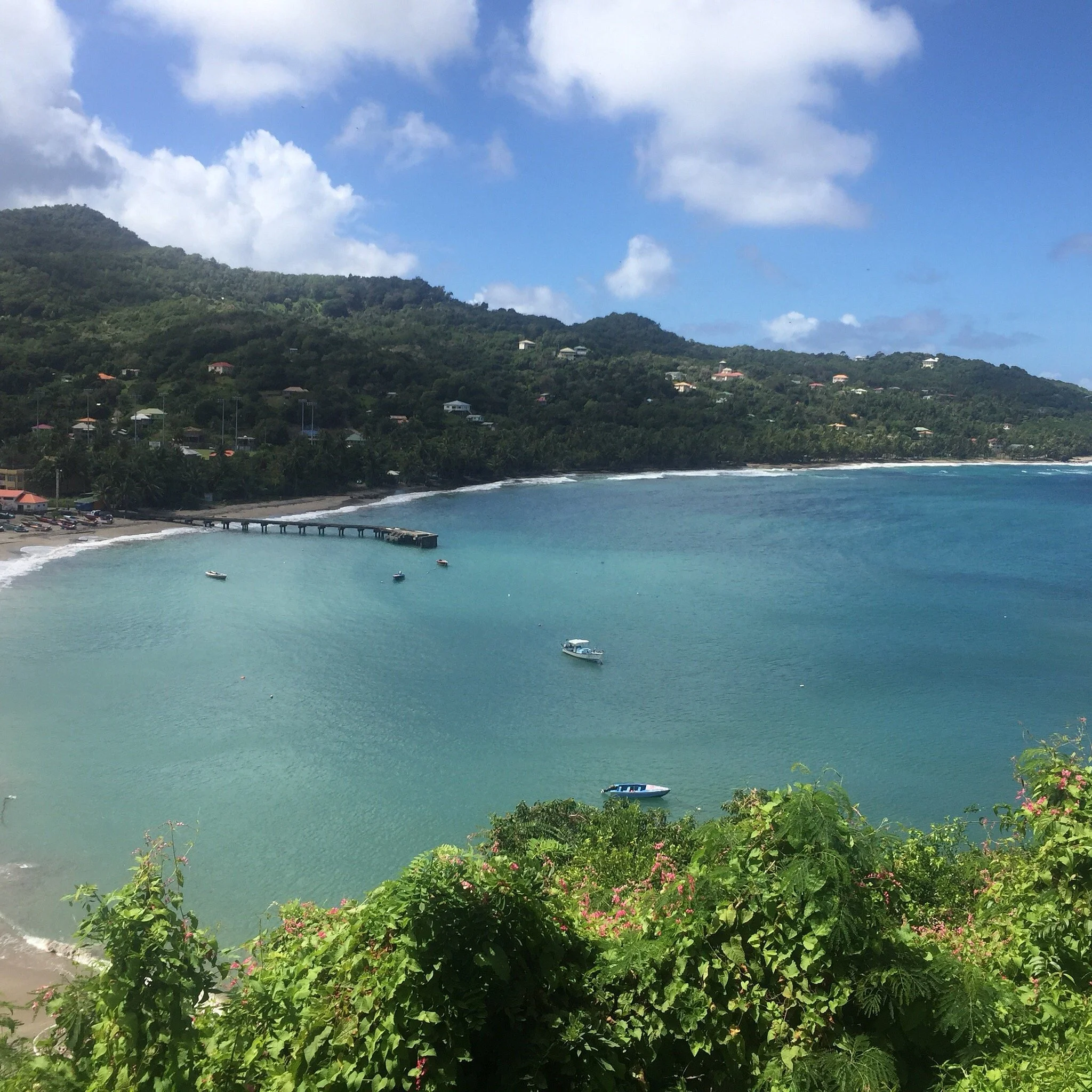 A scenic view of a bay with turquoise water, boats floating, a bridge or pier on the shore, lush green hills in the background, and a partly cloudy sky.