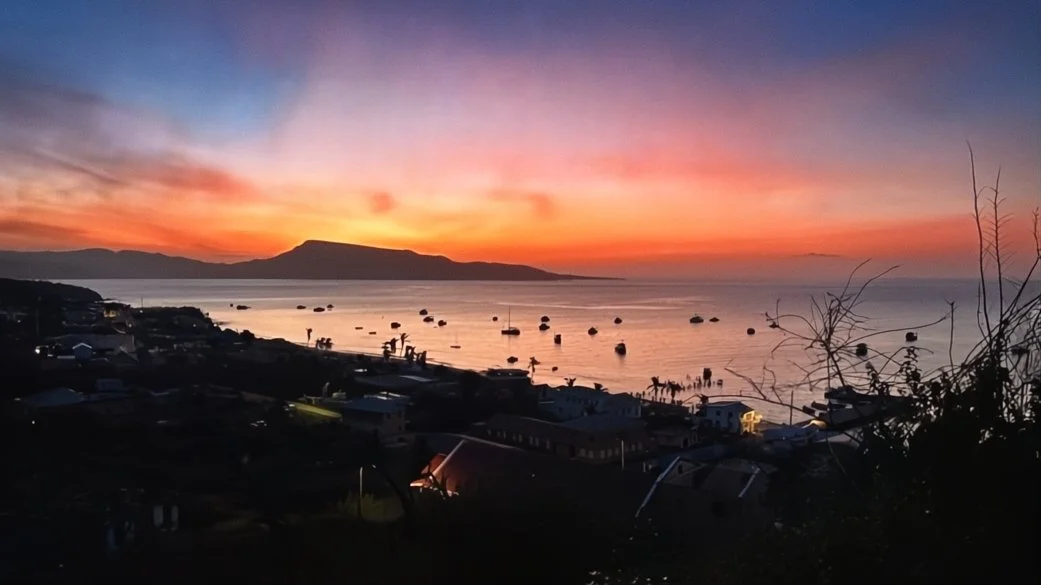 Sunset over a coastal town with a mountain in the distance, boats anchored in the water, and houses along the shoreline.
