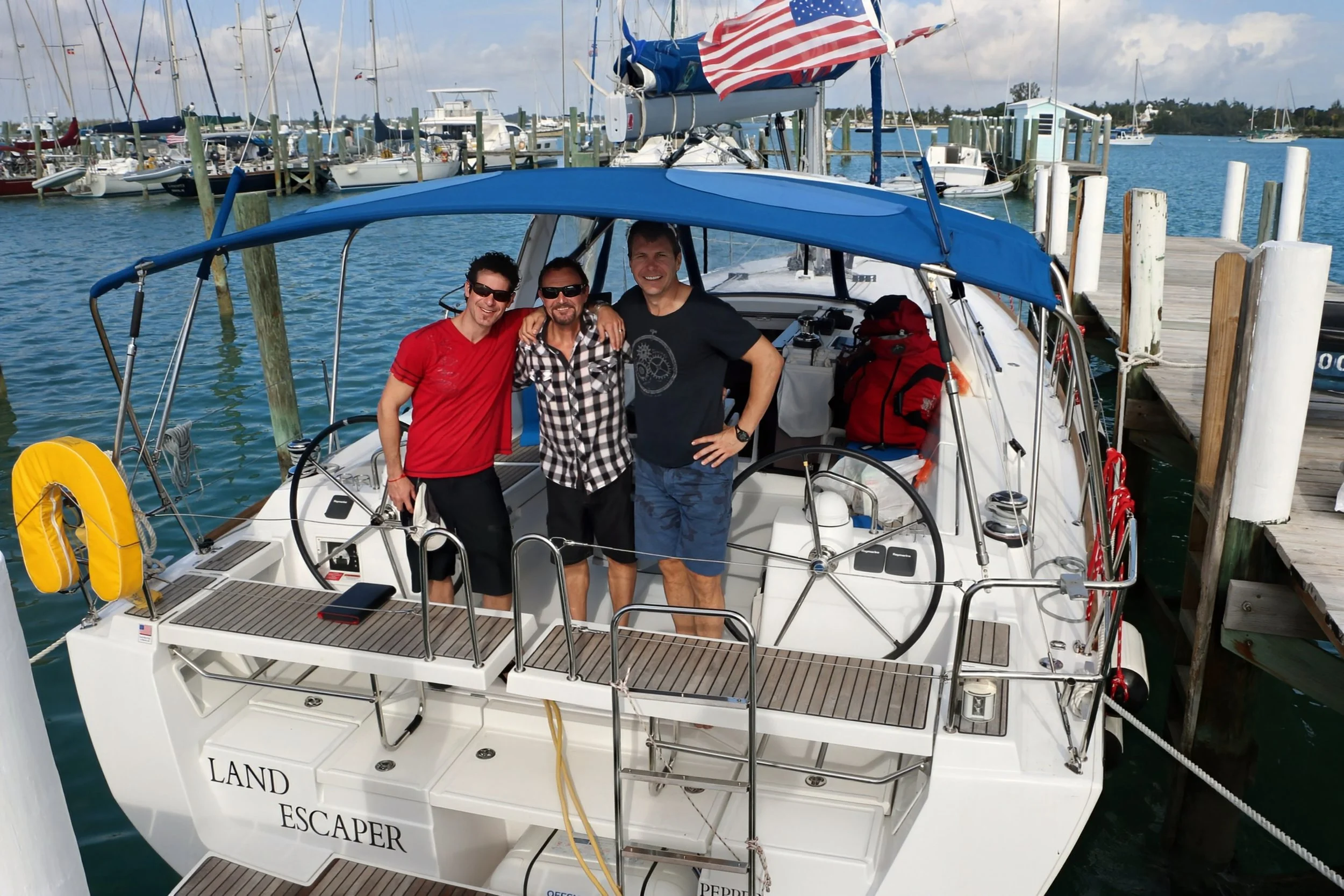 Three men standing on a white sailboat at a marina dock, smiling with arms around each other. The boat has a blue canopy, yellow life preserver, red backpacks, and a steering wheel. The dock and other boats are visible in the background.