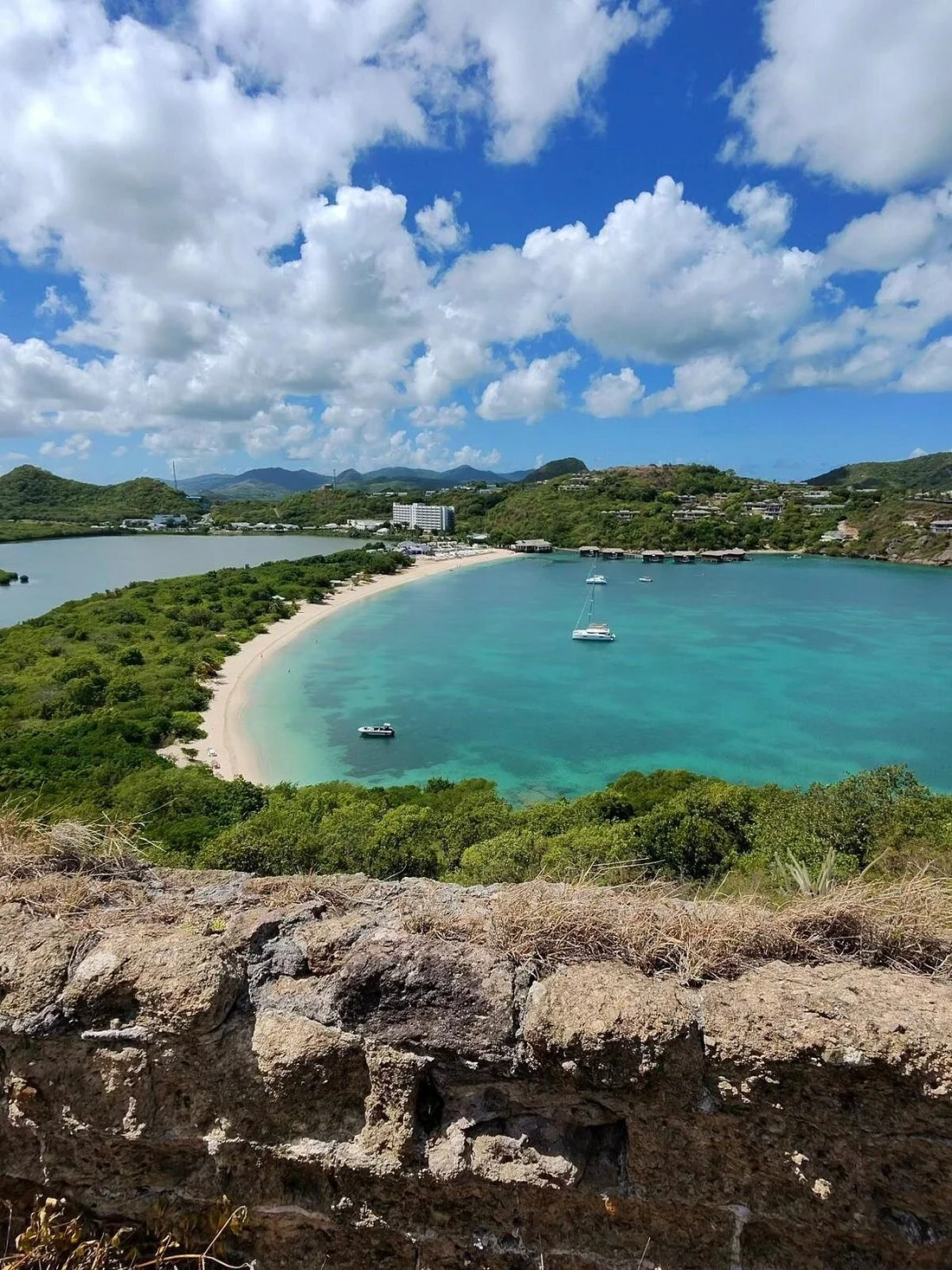 A tropical bay with clear turquoise water, white sandy beach, green foliage, and boats anchored. Hills and buildings can be seen in the background under a partly cloudy sky.
