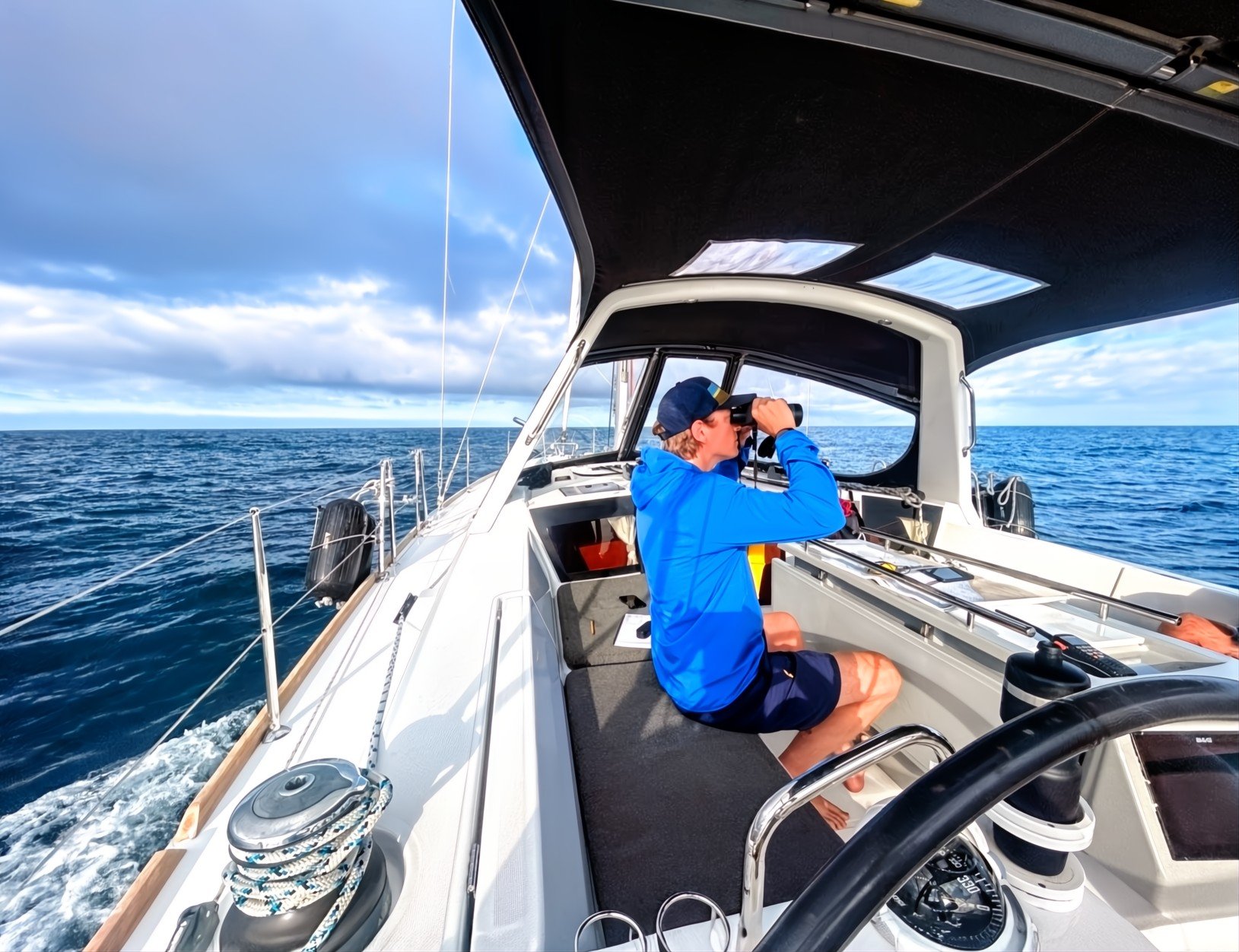 Person in a blue jacket and cap sitting on a sailboat looking through binoculars at the ocean under a partly cloudy sky.