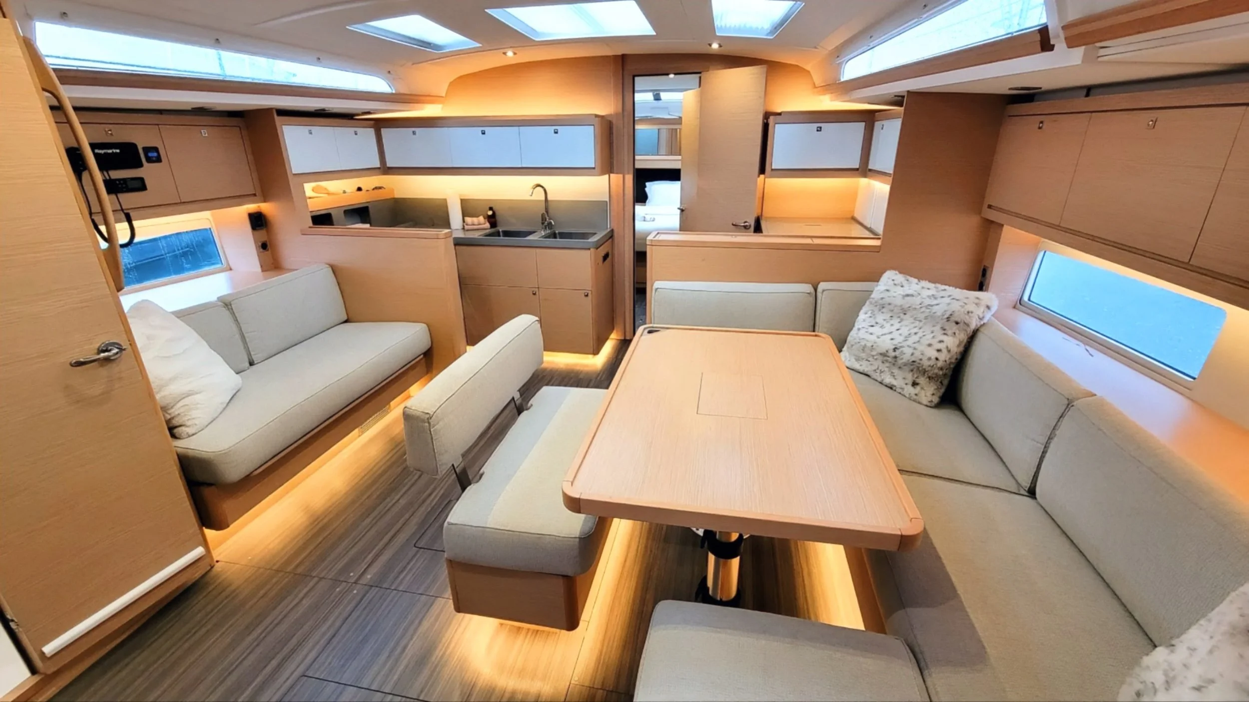 Interior of a modern Dufour 470 luxury yacht with beige sofas, a wooden table, a compact kitchen, wooden cabinets, and skylights.