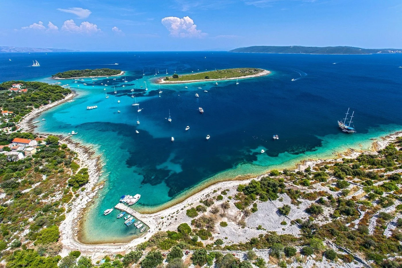 Aerial view of a coastal area with small boats in turquoise waters, surrounded by rocky shores and green vegetation, with island in the distance under a partly cloudy sky.