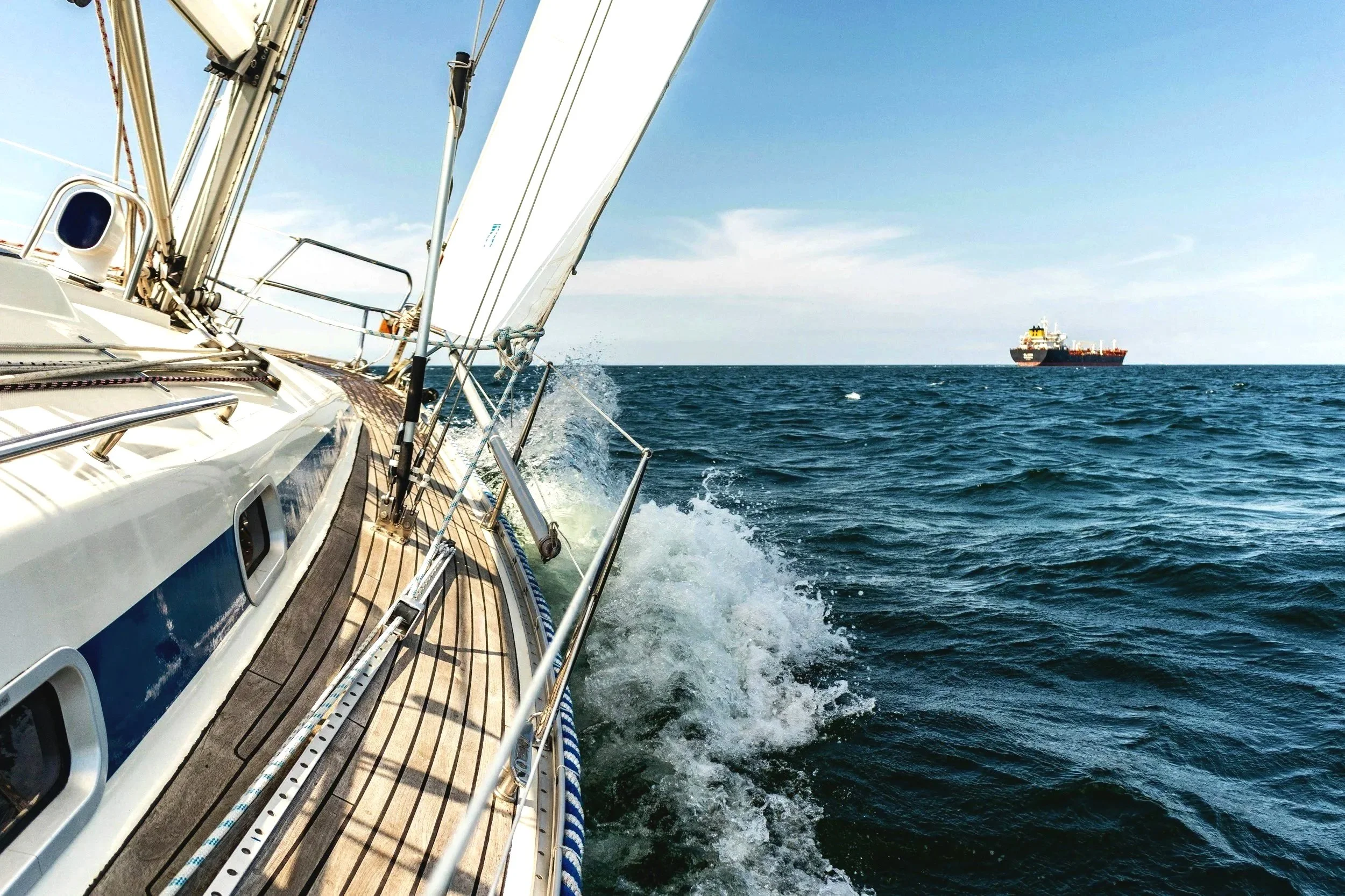 View from a sailboat looking toward the ocean with a large cargo ship in the distance, showing the boat's deck, rigging, and the waves splashing against the side.