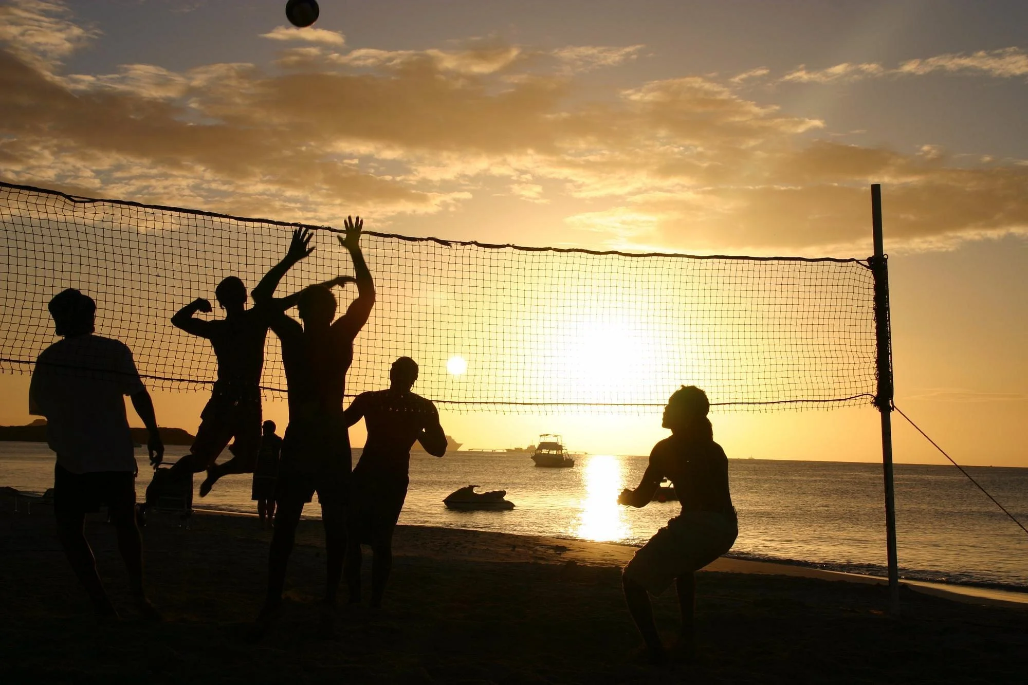 Silhouettes of people playing beach volleyball during sunset by the water.