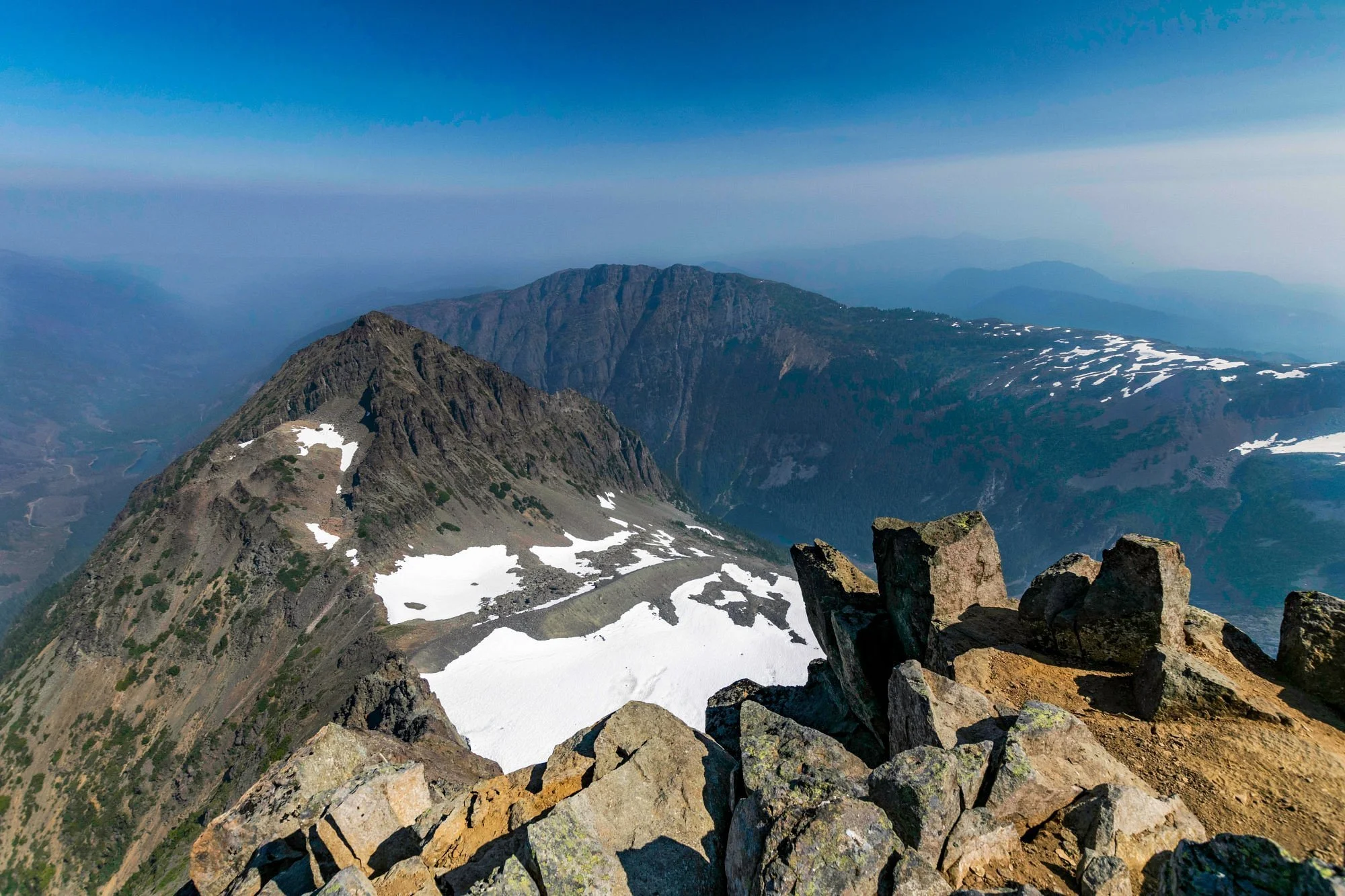 Mountain landscape with rocky foreground, snow patches on slopes, and distant mountain ranges under a clear blue sky.