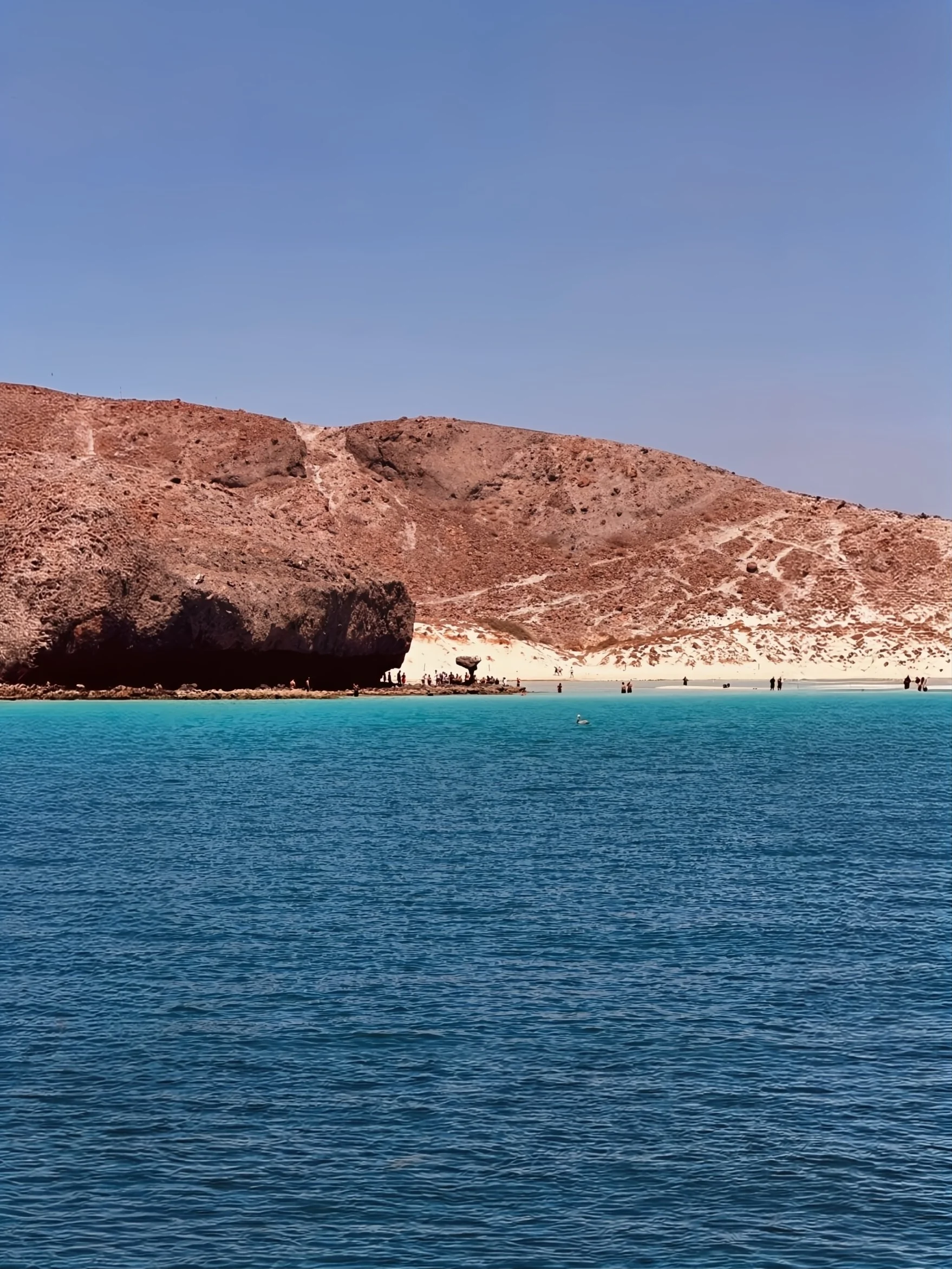 A scenic view of a turquoise lake with rocky, desert-like mountains in the background and a clear blue sky overhead.