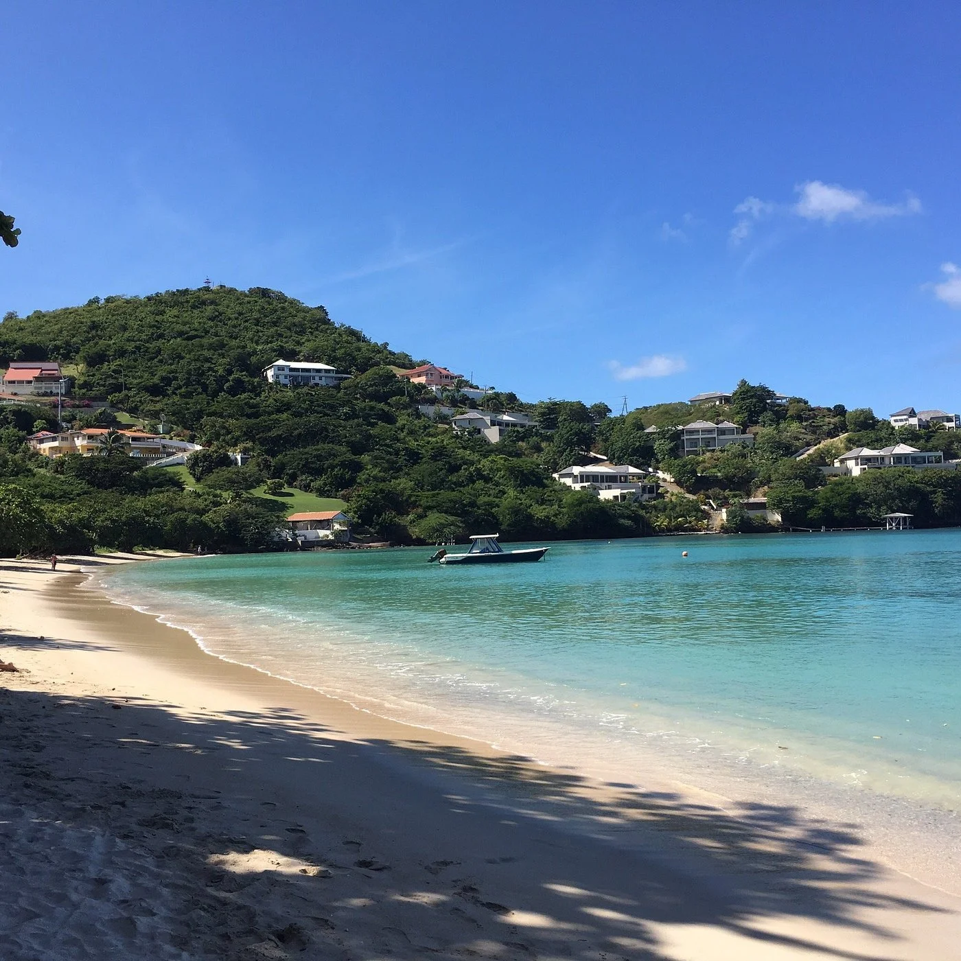 A beach with white sand, clear turquoise water, and a boat anchored near the shore. Hills with houses are visible in the background under a blue sky with a few clouds.