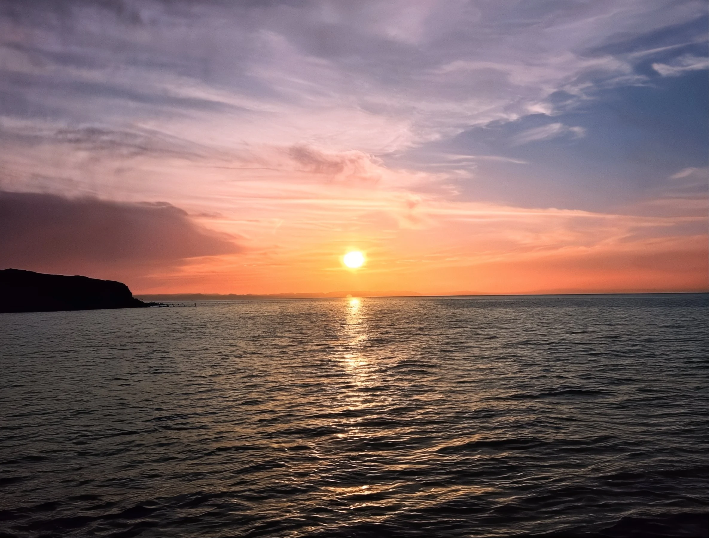 A peaceful sunset over the ocean with a partly cloudy sky and a dark silhouette of land on the left.