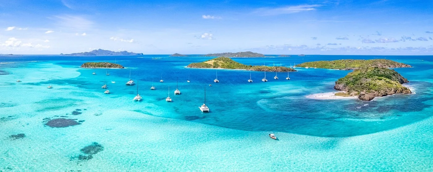 An aerial view of a tropical island with turquoise waters, several sailboats anchored, and lush green islands in the background under a blue sky with some clouds.