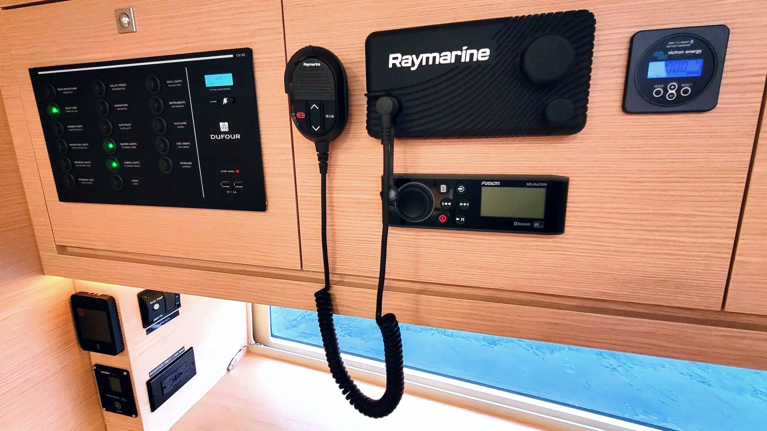 View of a wooden panel with various electronic and communication devices, including a control panel with labeled switches and lights, a Raymarine marine device, a Fusion radio, a remote control, a digital temperature controller, and a mounted microph