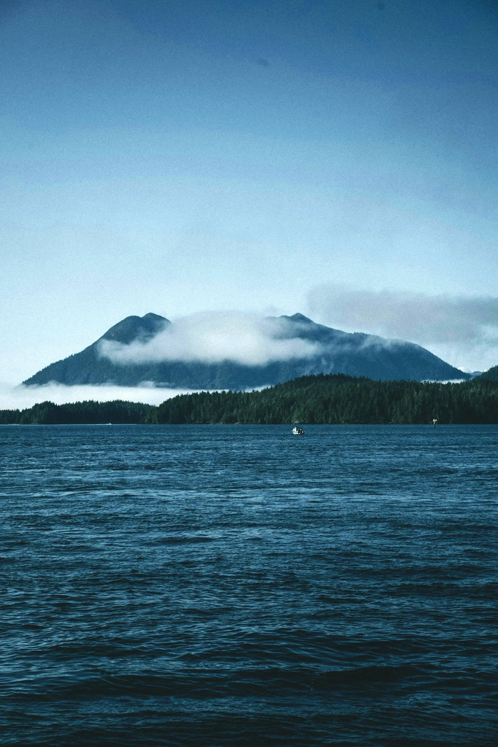 A large body of water with small ripples, forested land in the background, and a mountain with clouds surrounding its peaks under a blue sky.