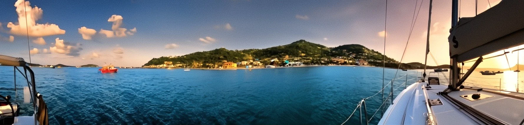 View from a sailboat looking across a calm bay with small boats, a hillside dotted with houses, and a partly cloudy sky during sunset.