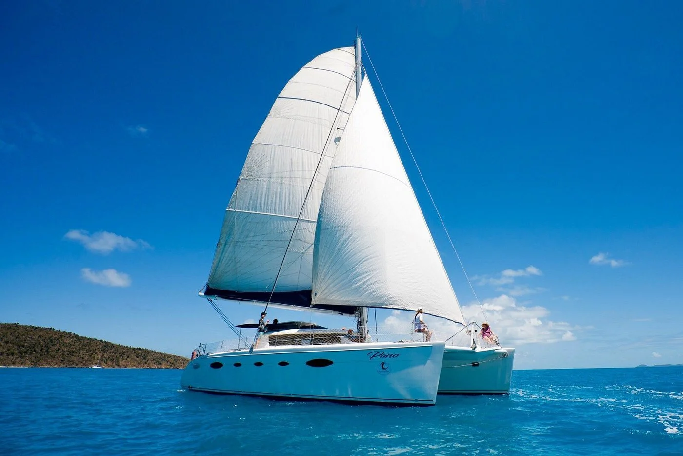 A white sailboat with two masts sailing on blue water with a hilly landmass in the background, under a clear blue sky with a few clouds.