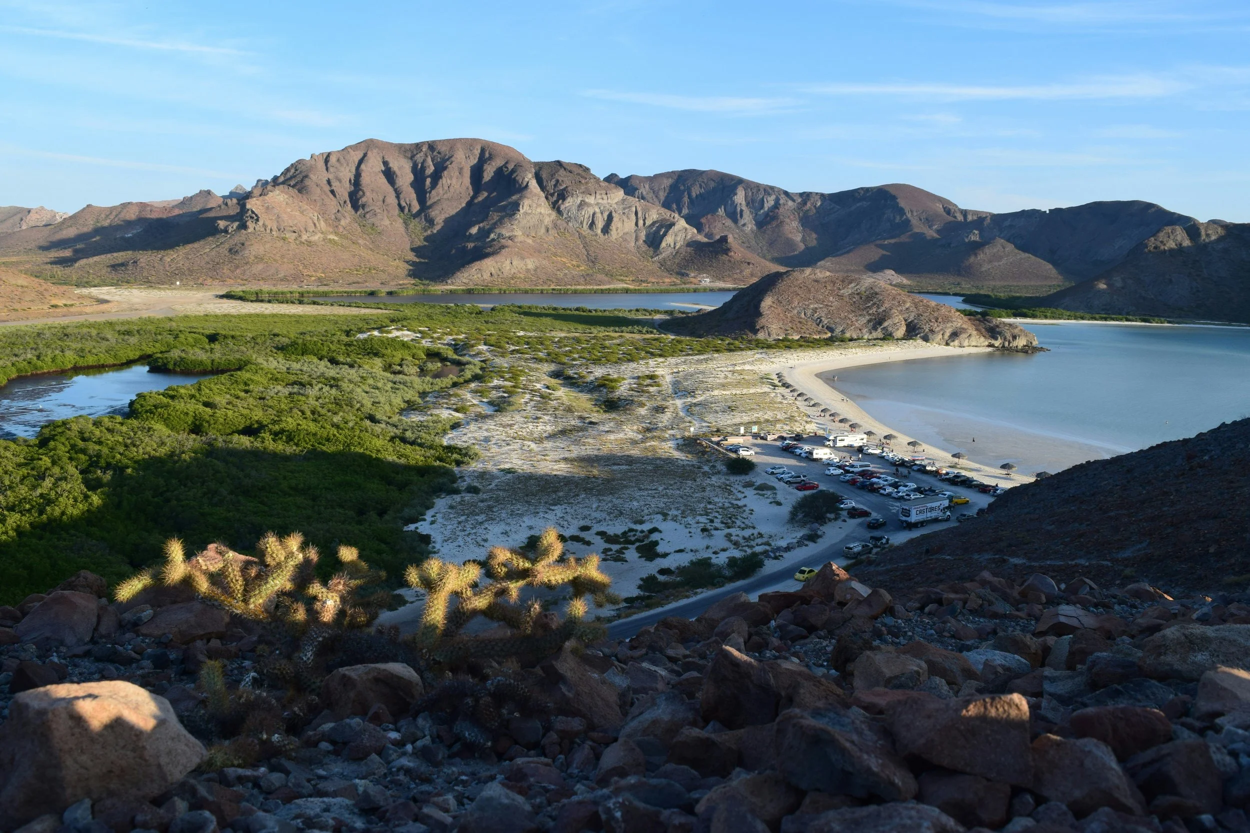 A landscape view of a winding river near a sandy beach, with a parking lot full of cars, surrounded by mountains and green vegetation under a blue sky.
