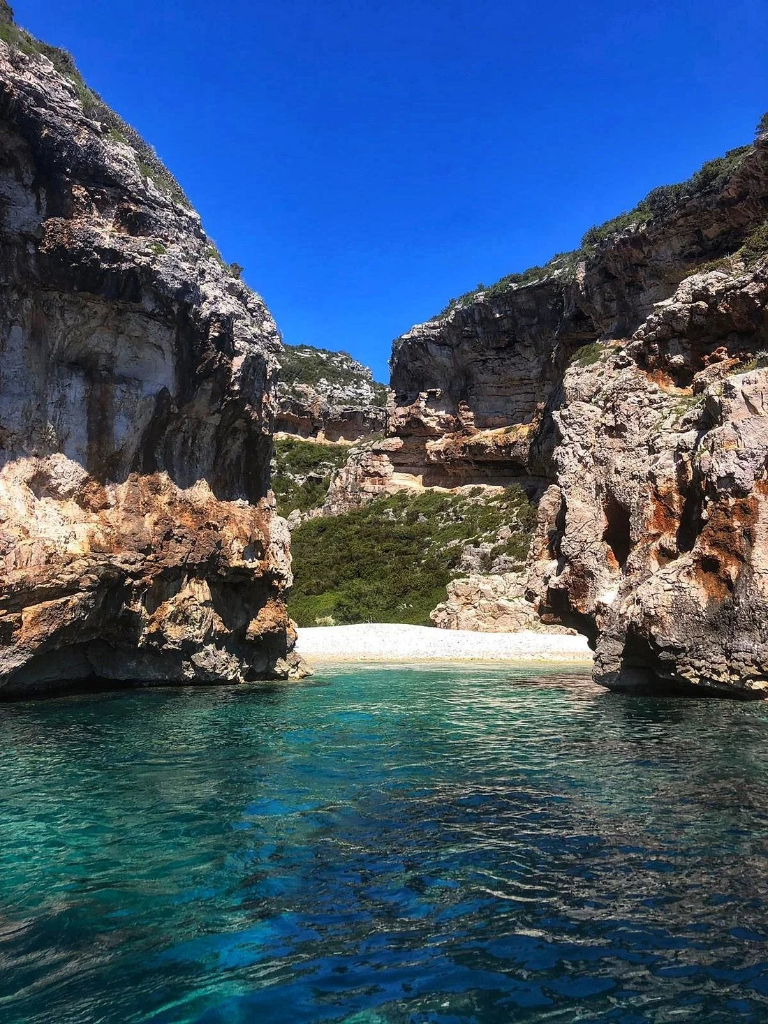 A scenic view of a cove with clear turquoise water, surrounded by tall rocky cliffs under a bright blue sky.
