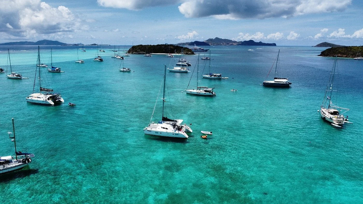 Aerial view of sailboats and yachts anchored in turquoise water near islands under a partly cloudy sky.