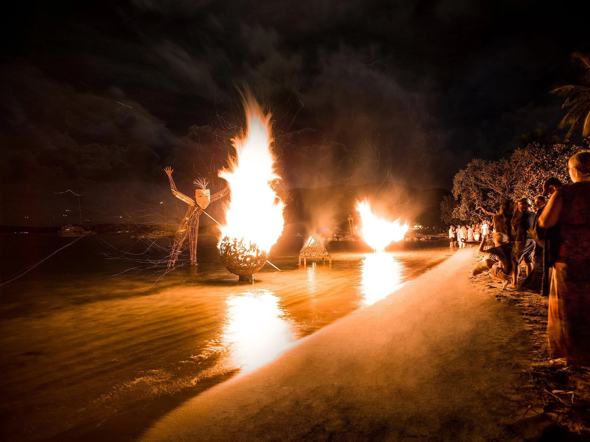 A nighttime scene at a beach with large bonfires and a crowd of people gathered along the shoreline. A large puppet or effigy with outstretched arms and a mask is near the bonfire on the left. The sky is dark, and there are some trees visible on the 