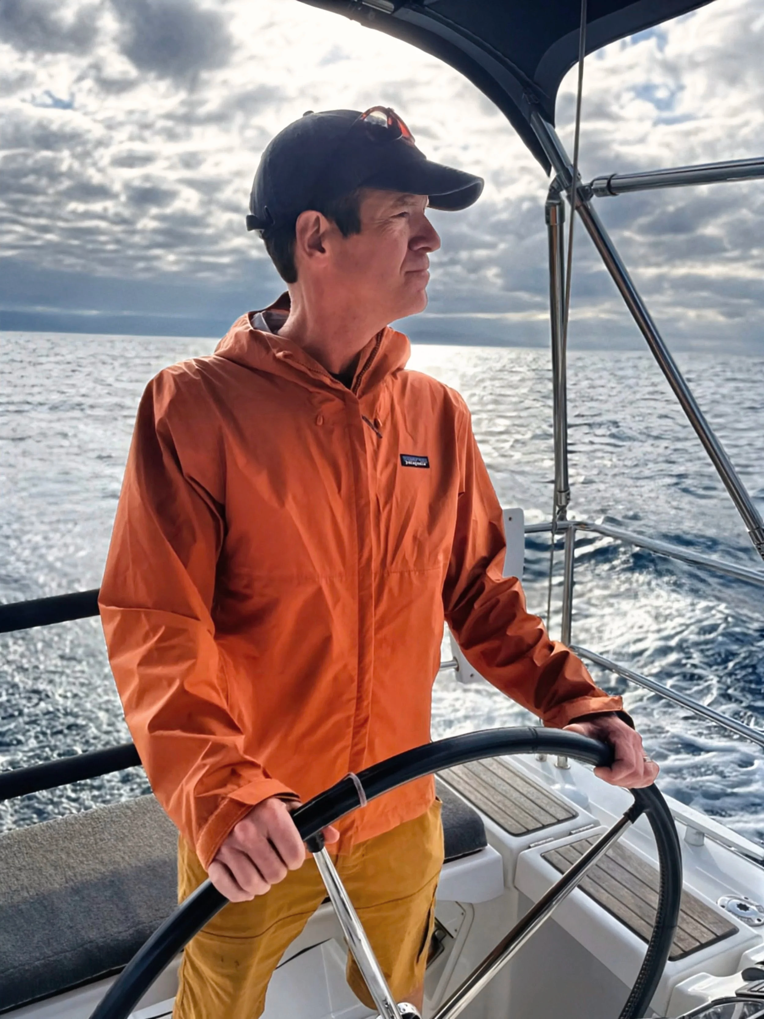 A man wearing an orange jacket and a black cap steering a boat on the ocean. The sky is cloudy with sunlight breaking through.