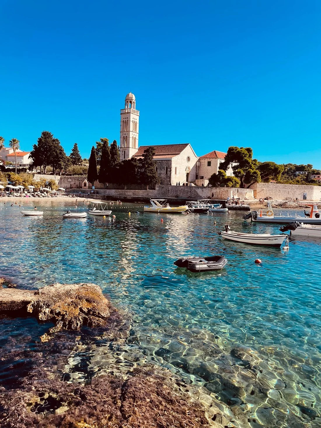 Scenic view of a coastal town with boats anchored in clear water, historic stone church with a tall bell tower, and lush greenery under a bright blue sky.