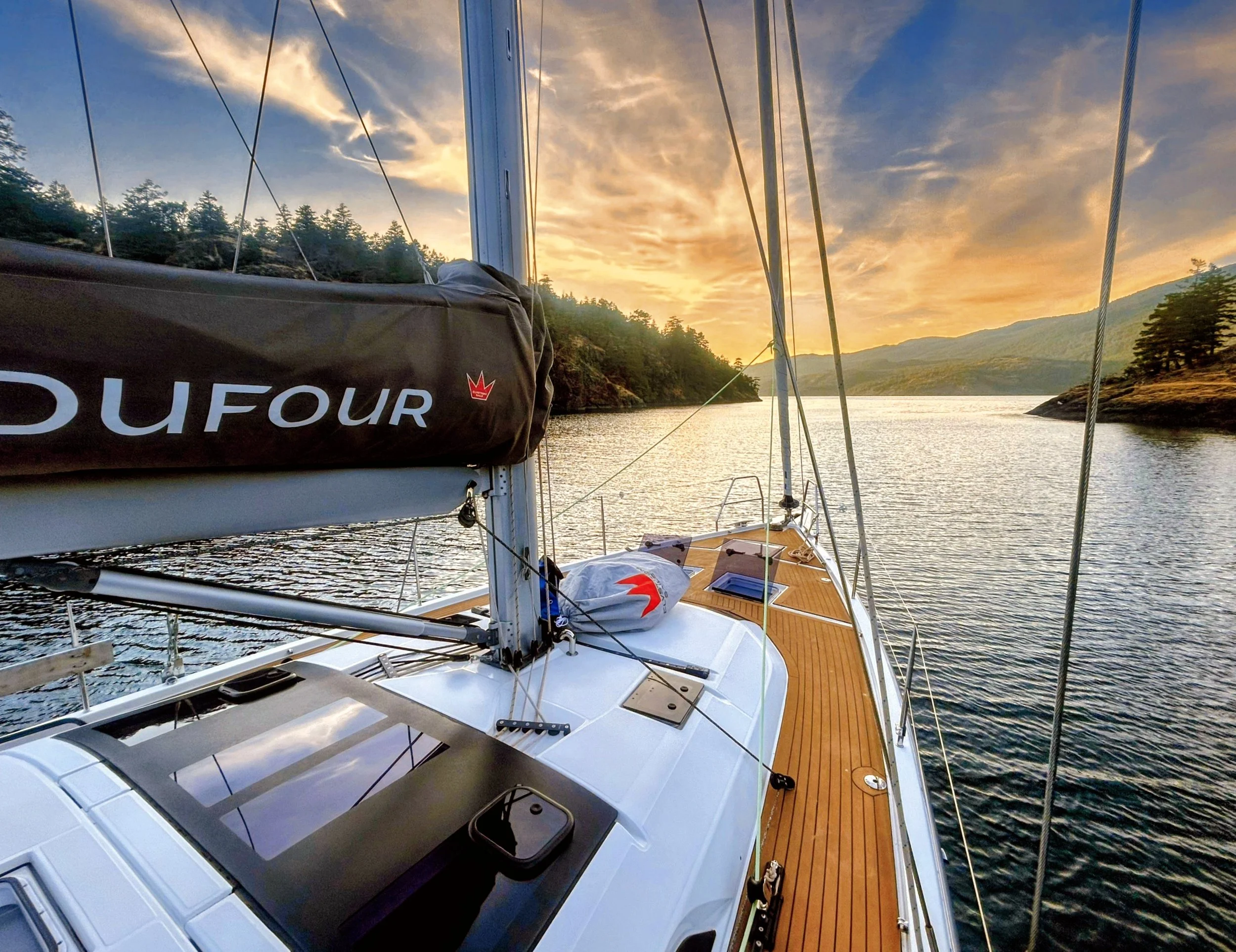 View from a sailboat's deck overlooking a river at sunset, with surrounding trees and distant mountains.
