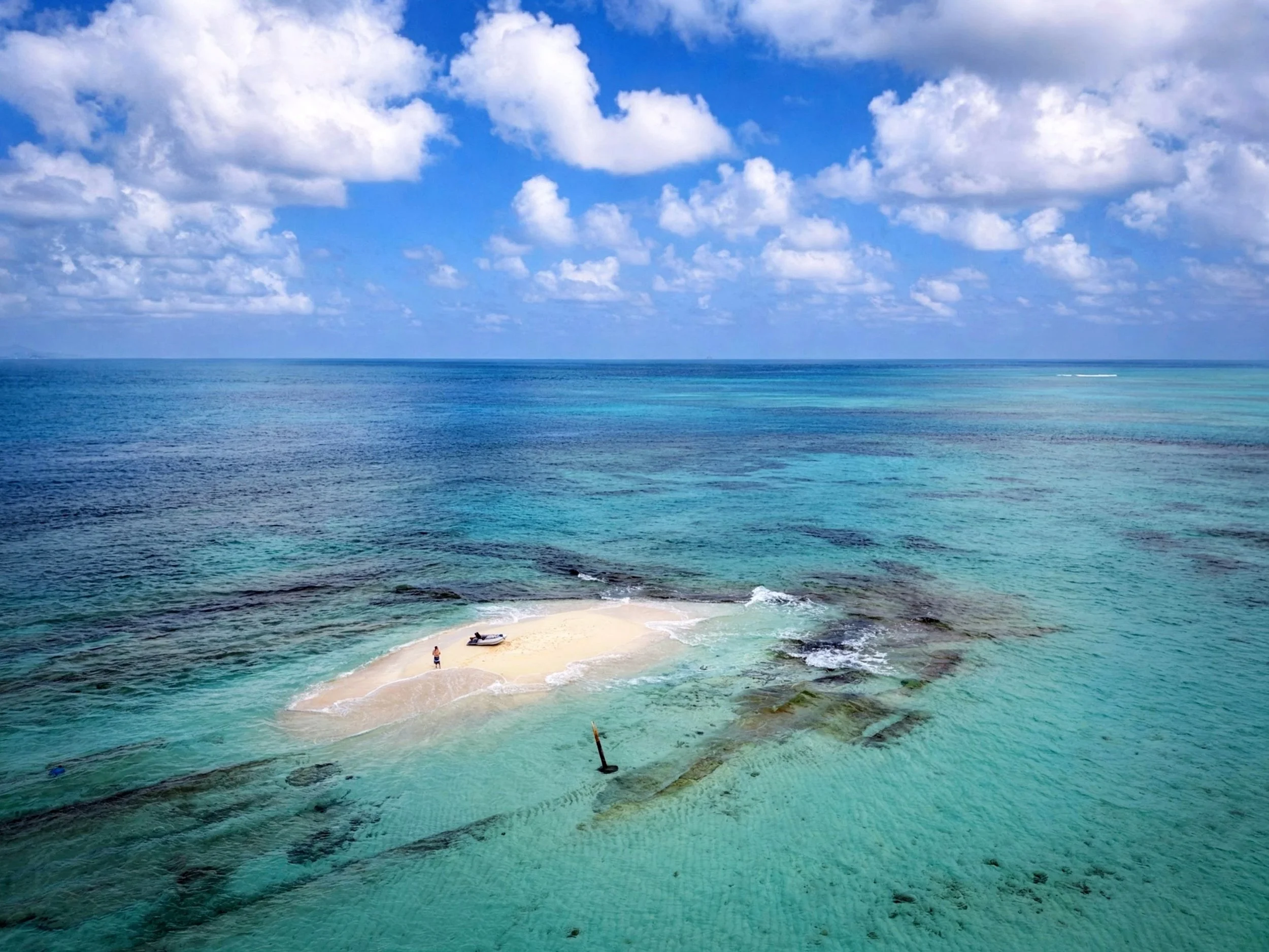A small sandy island with a boat and a person on it, surrounded by turquoise ocean water and underwater rocks, under a partly cloudy sky.