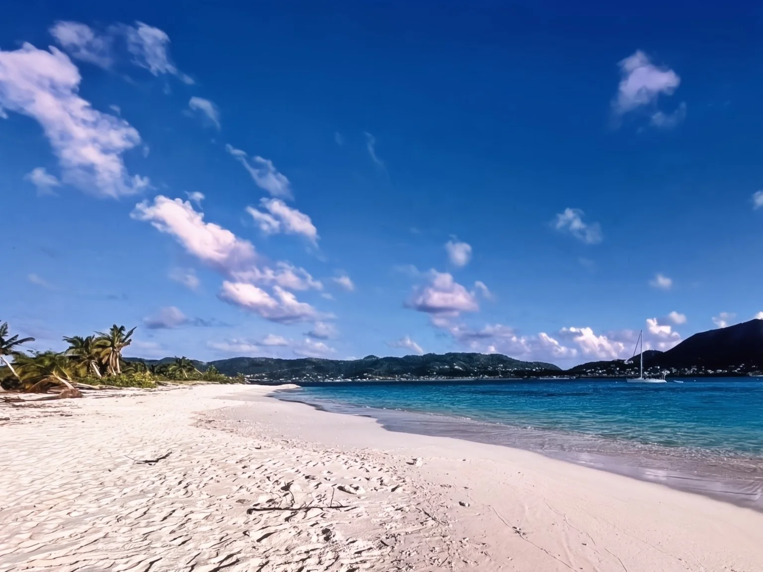 Tropical beach with white sand, palm trees, calm turquoise water, and a sailboat under a blue sky with scattered clouds.