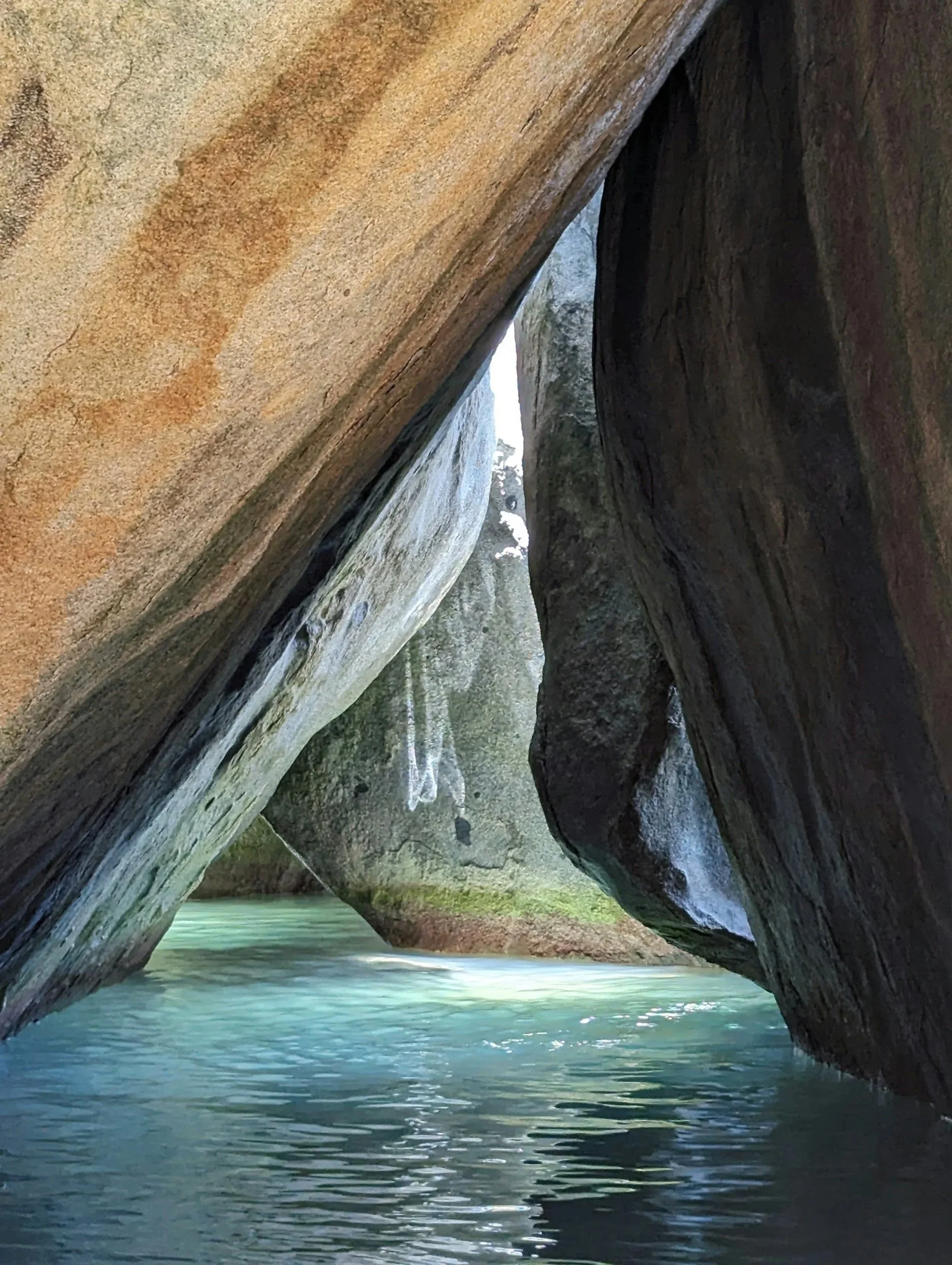 A narrow waterway with large, smooth rock formations on either side, some with visible water streaks and moss, and sunlight reflecting off the water's surface.