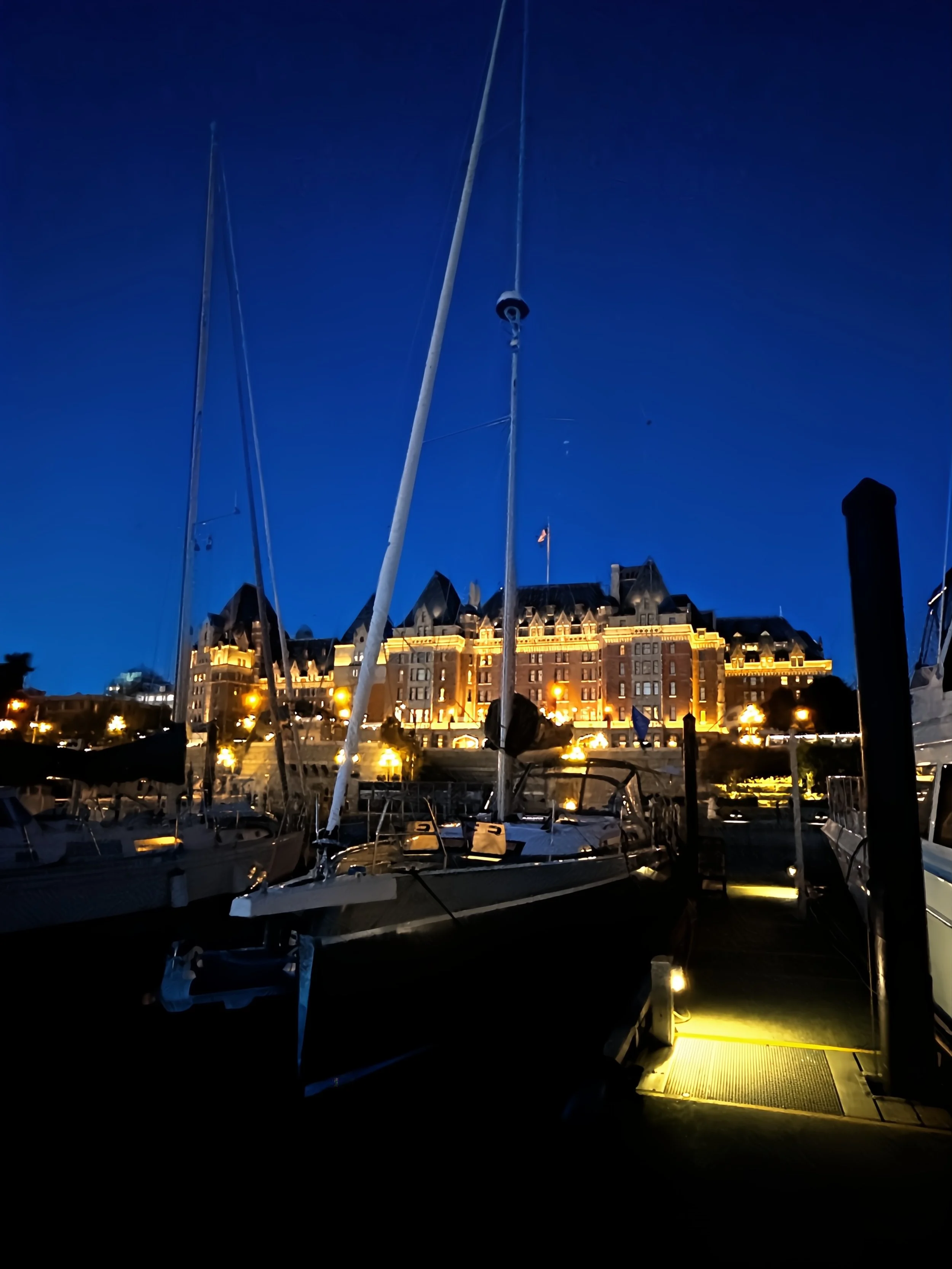 Boats docked at a marina at night, with a large brightly lit building in the background under a dark blue sky.