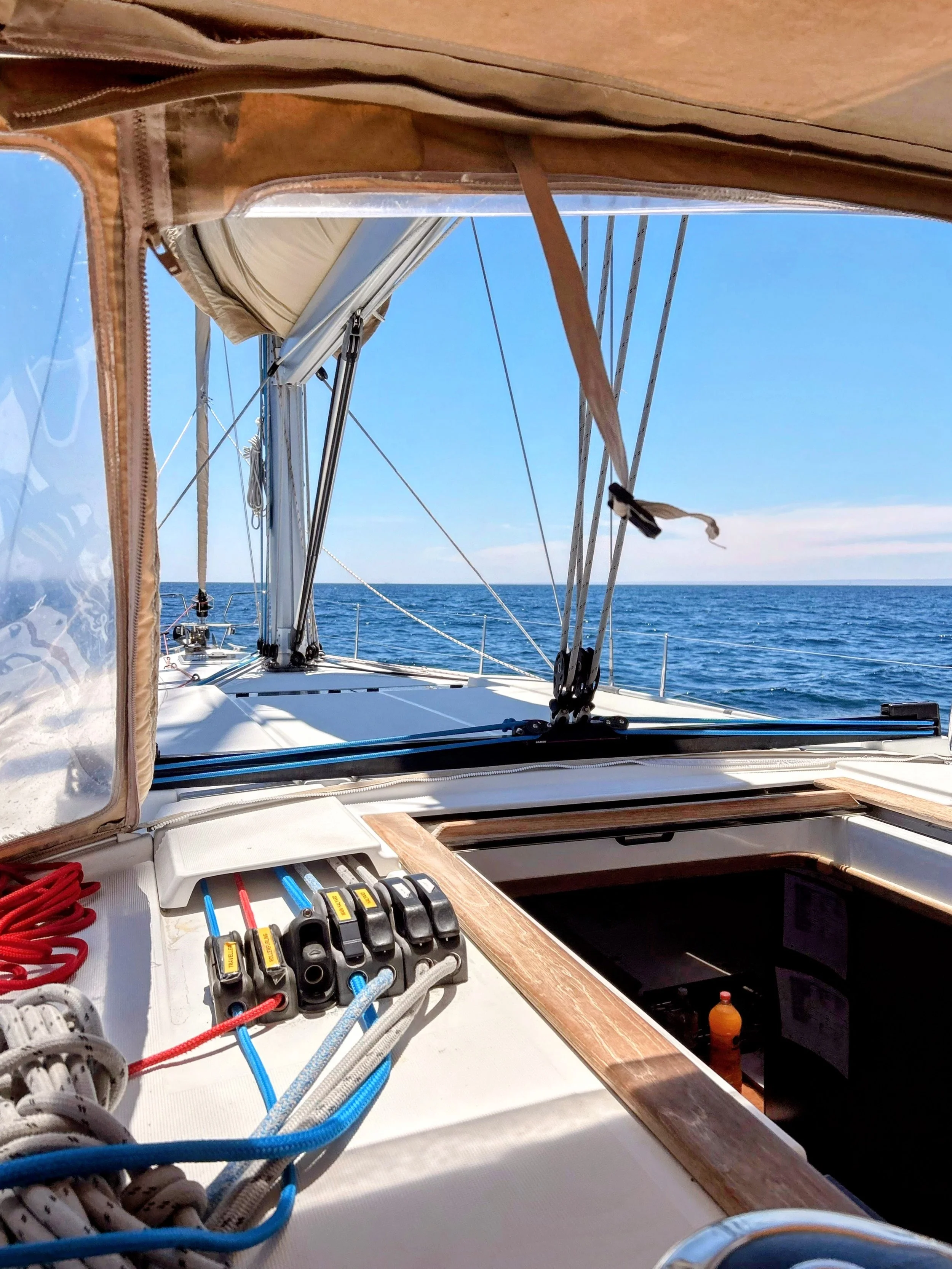 View from a sailboat deck looking out at the open ocean with a clear blue sky, showing sailing rigging, ropes, and part of the cockpit with electronic instruments.